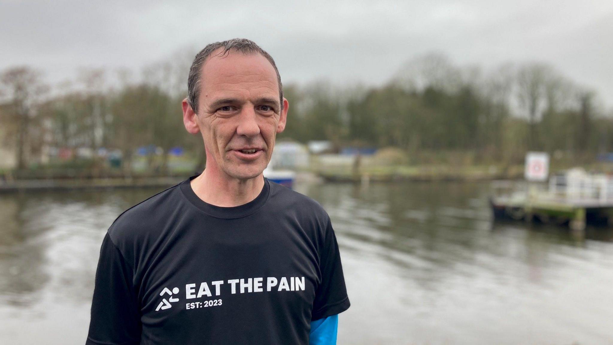 Mark Richards wearing a T-shirt that says 'eat the pain' stood at Lough Neagh after completing Antrim parkrun