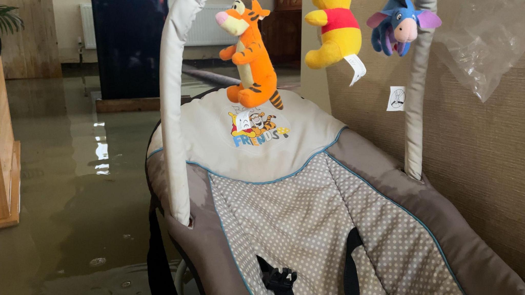 At least two inches of water can be seen in the interior of a living room with a baby seat in the foreground with toy characters from Winnie the Pooh hanging above it.