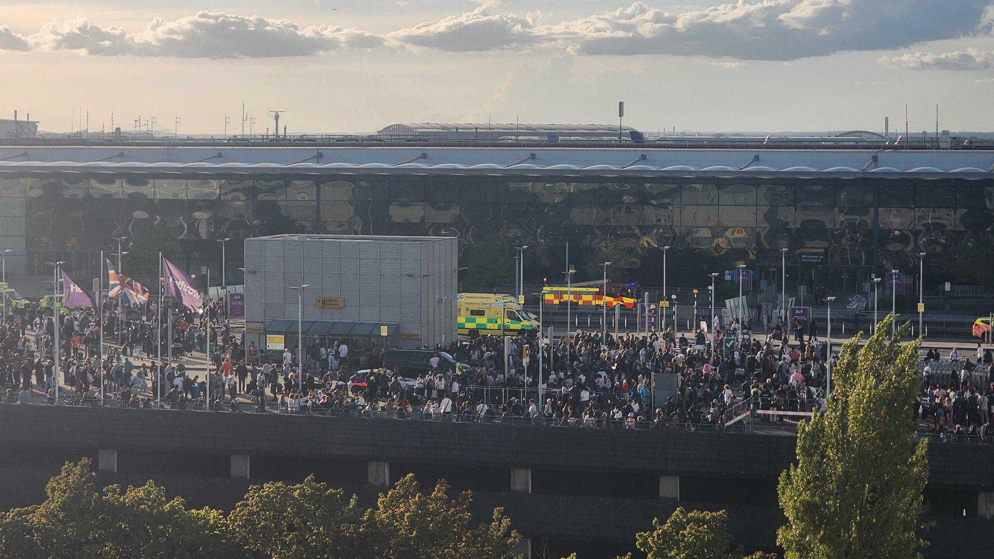 Large crowd of passengers gathered outside Heathrow Terminal 4 alongside multiple ambulances and emergency vehicles, after the building was evacuated during a safety incident.