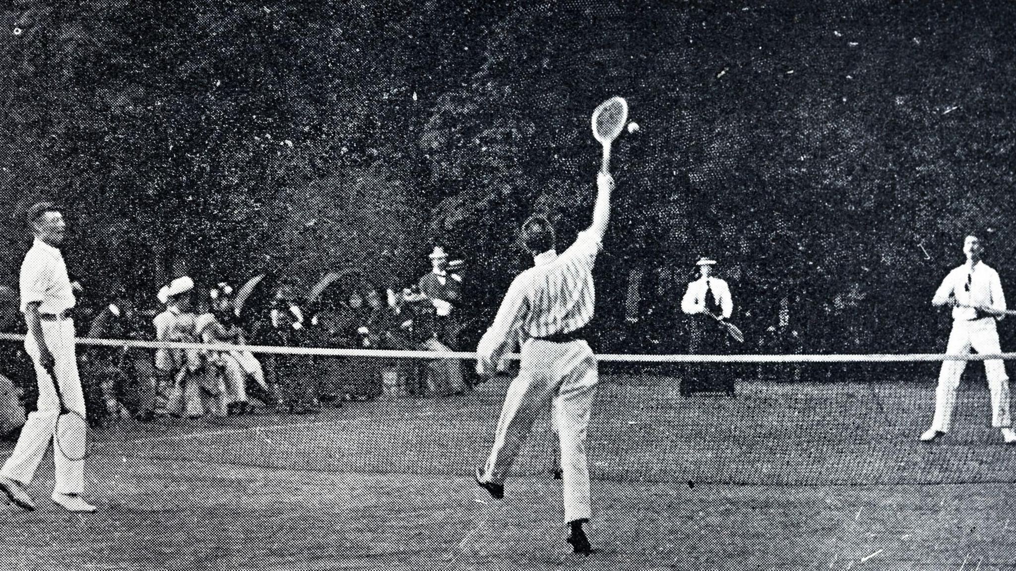 A black and white image of a19th Century lawn tennis match. Three men and a woman hold wooden rackets, with a seated crowd sat by the sidelines.