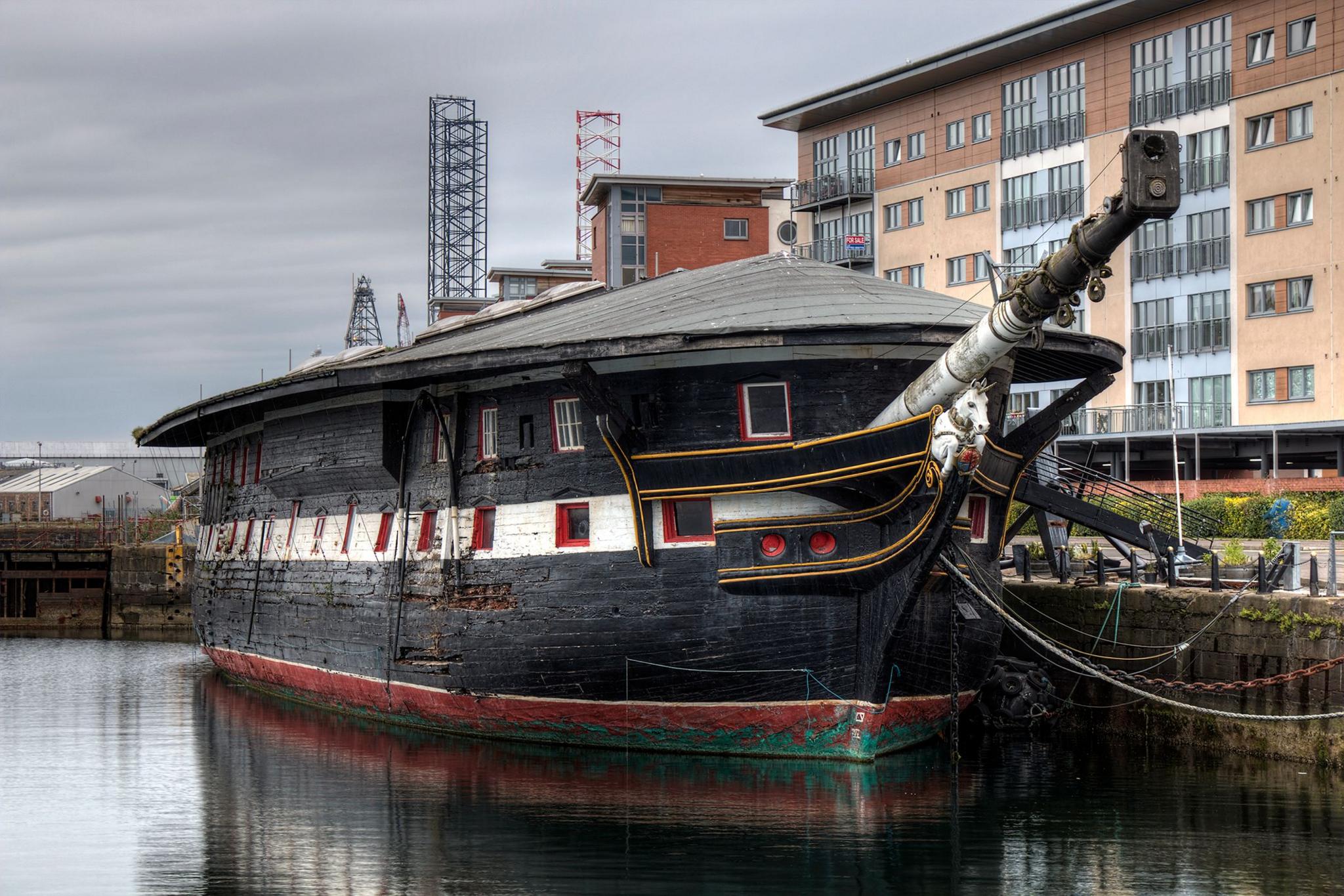 A large black ship with white and red detailing ship dating from the 1800s sits in a dock, with a modern apartment building in the background