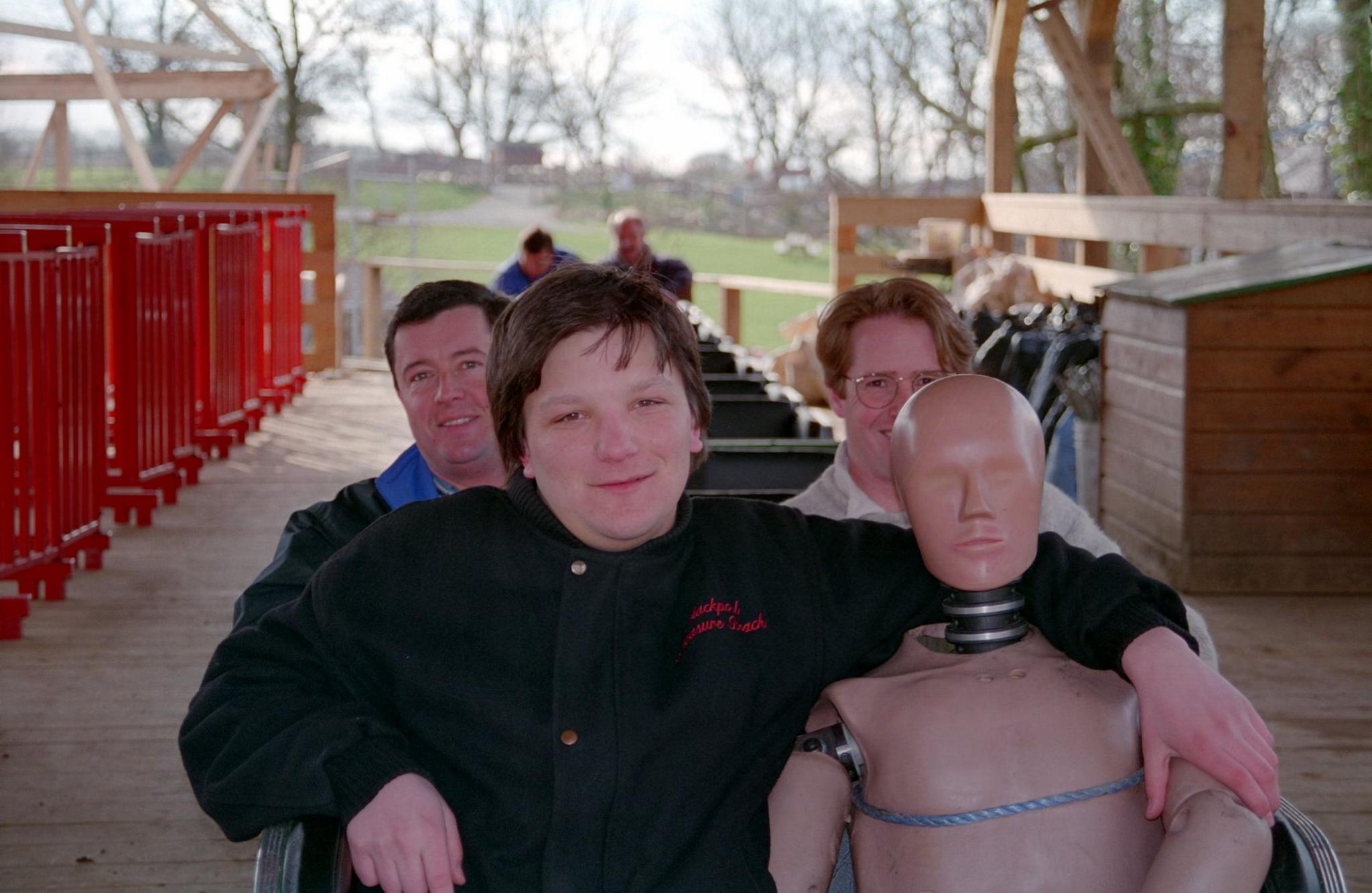 Justin Garvanovic sat in the front row of the Megafobia ride at Oakwood with his arm around a test dummy figurine. There are two men in the row behind. In the background more people are in the carts at the tail end of the rollercoaster with the green setting of Oakwood with trees and fields behind them.