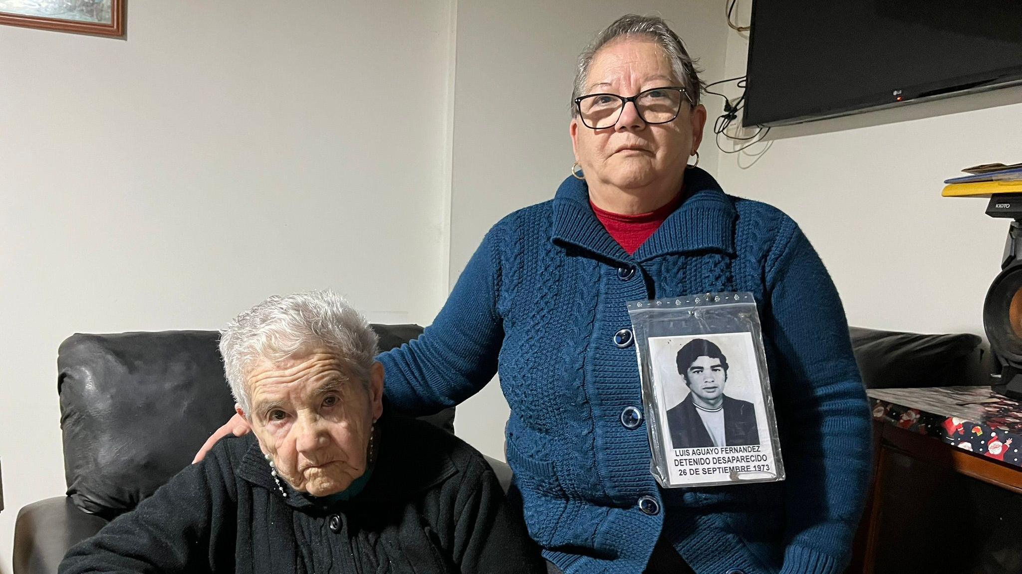 Ana Aguayo's mother, wearing a black cardigan and sitting in a leather chair, looks into the camera as her daughter has her hand placed on her shoulder. 
Ana has the photo of her brother pinner to her cardigan. 