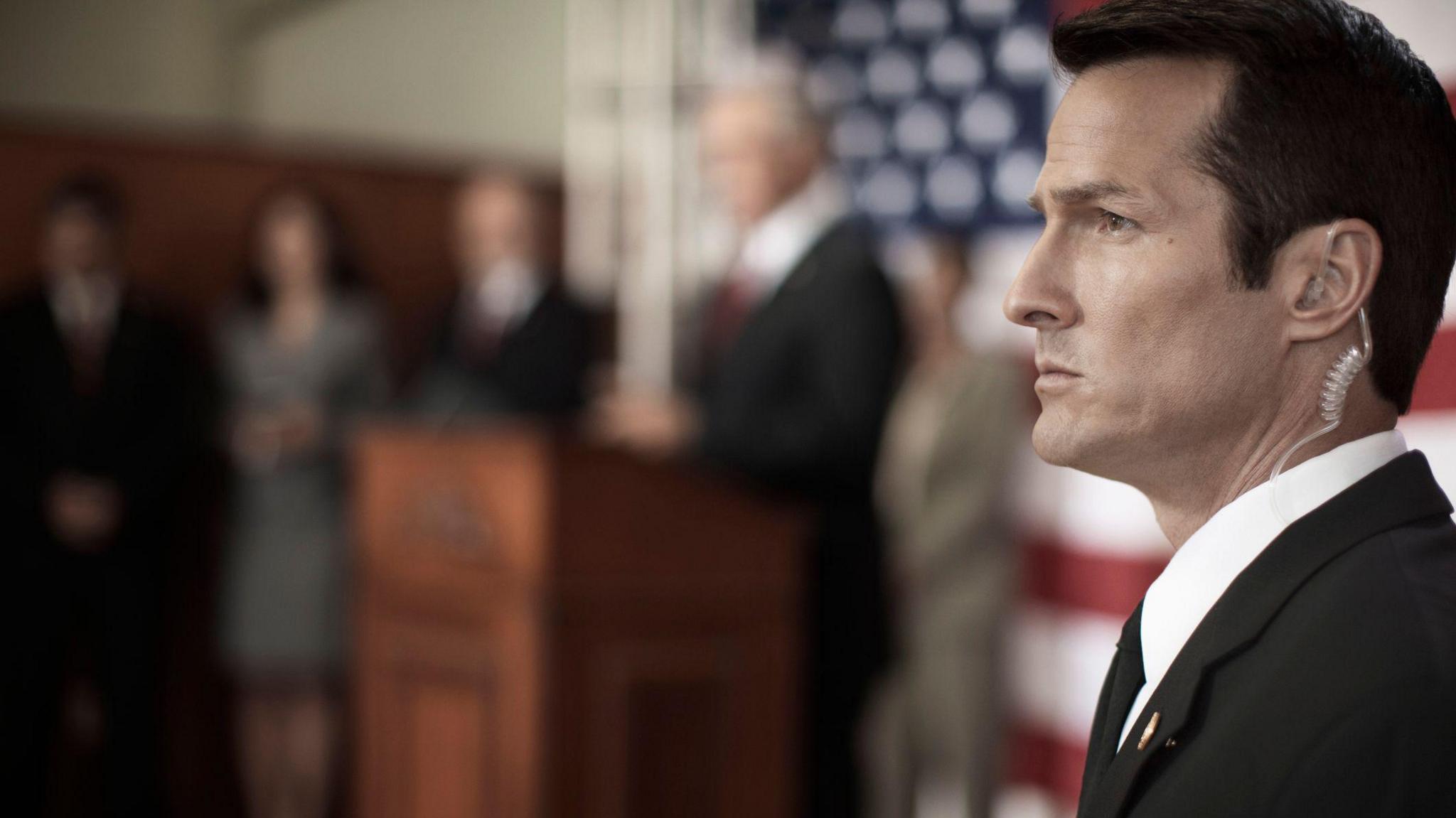 Secret service agent with earpiece in while someone does a speech in the background