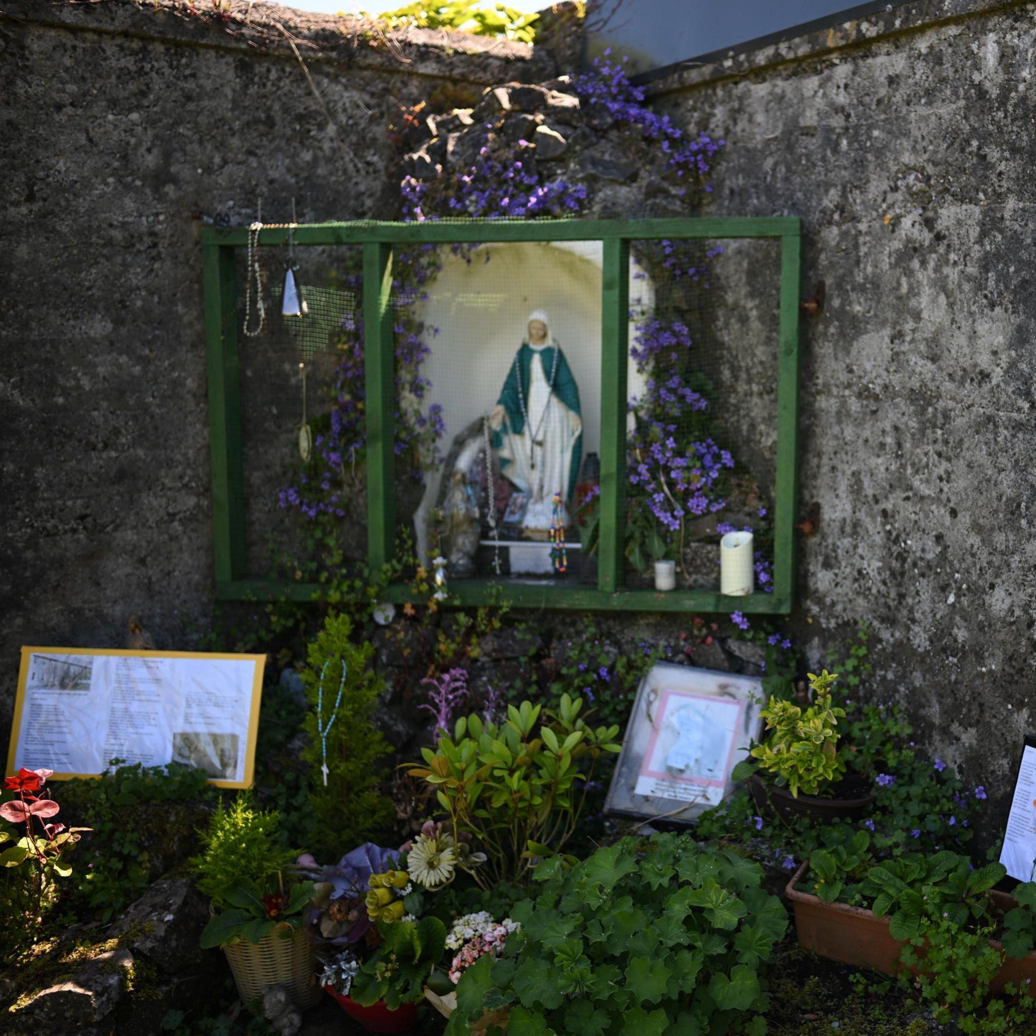 Two high stone walls intersect at right angles in a garden - in the corner of the angle, sits a grotto centred on a statue of Virgin Mary. It is surrounded by flowers and shrubs. There are some candles on a shelf near the statue's feet. There are also messages underneath the statue that have been left by people and, to the right, a white board on which a message of remembrance is written