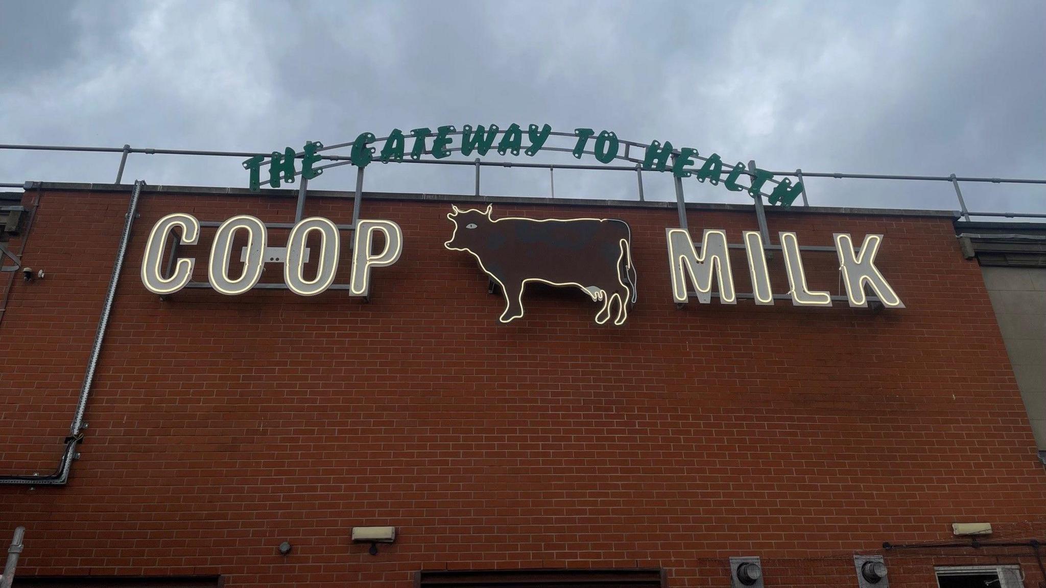 A sign with green lettering saying "The Gateway to Health" and white lettering underneath saying "Co-op Milk" with a cow figure in the middle. It is on the side of a brick building 