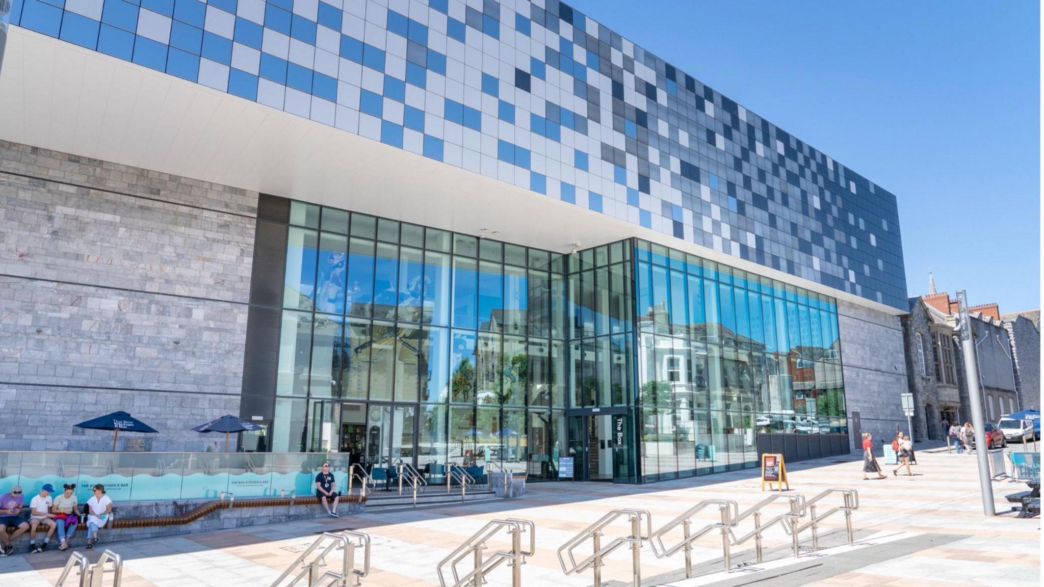 The image shows the exterior of the Box Museum and Art Gallery in Plymouth on a bright sunny day. It is a modern building at the rear which is shown, with very large glass windows covering much of the side. A pedestrianised area lies in front.