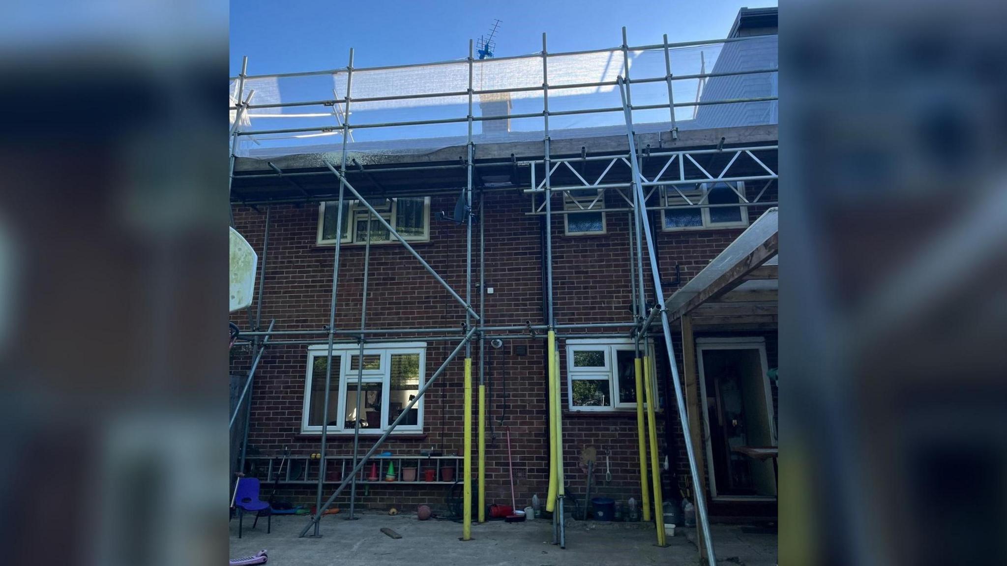 A mid-terrace brick property with scaffolding covering the entire front. There is a ladder at the front. Yellow foam surrounds some of the scaffolding poles.