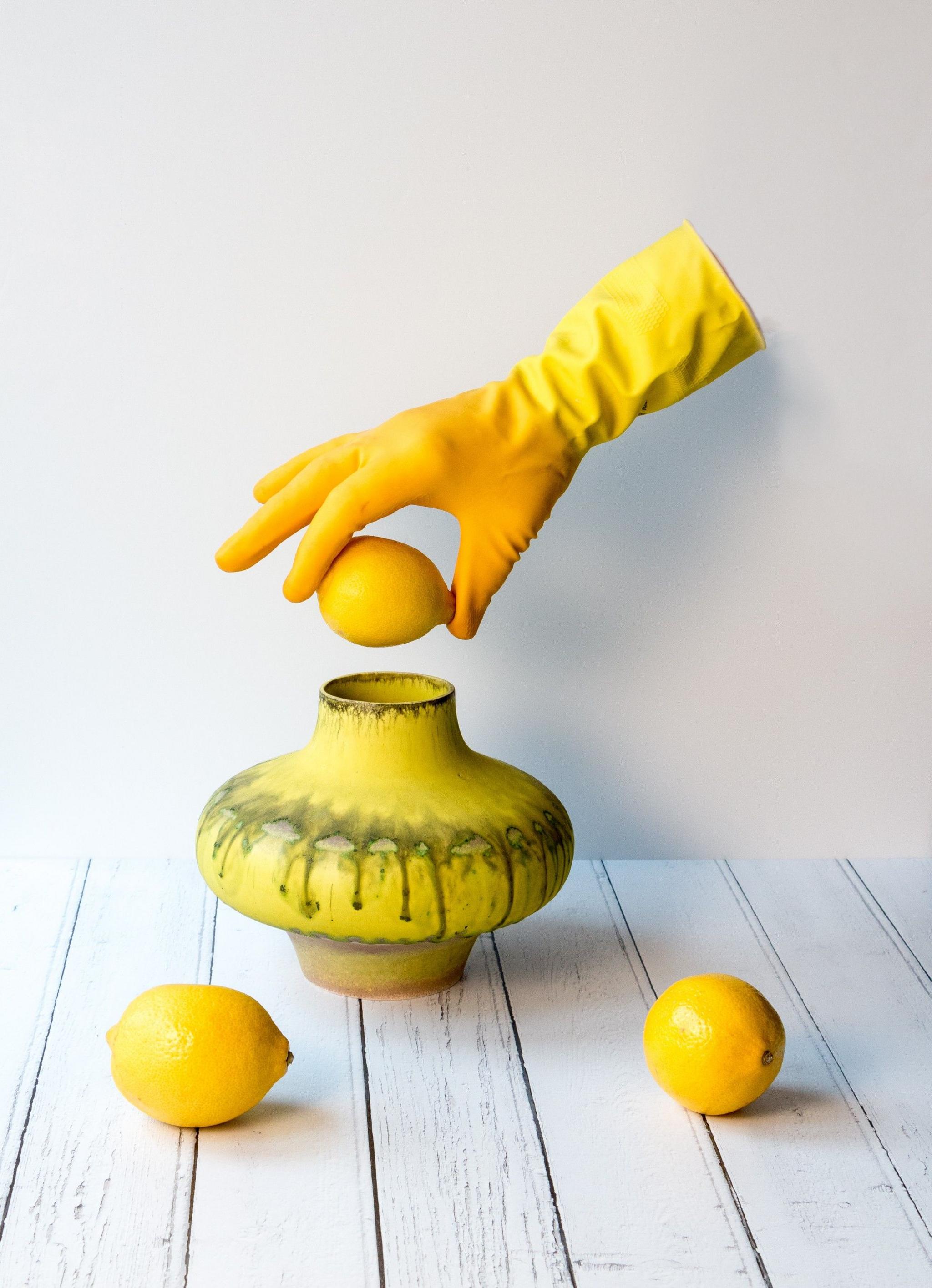 A yellow gloved hand, suspended in the air without being attached to a body, holds a lemon above a yellow vase. Two more lemons sit on a table in front of the vase.