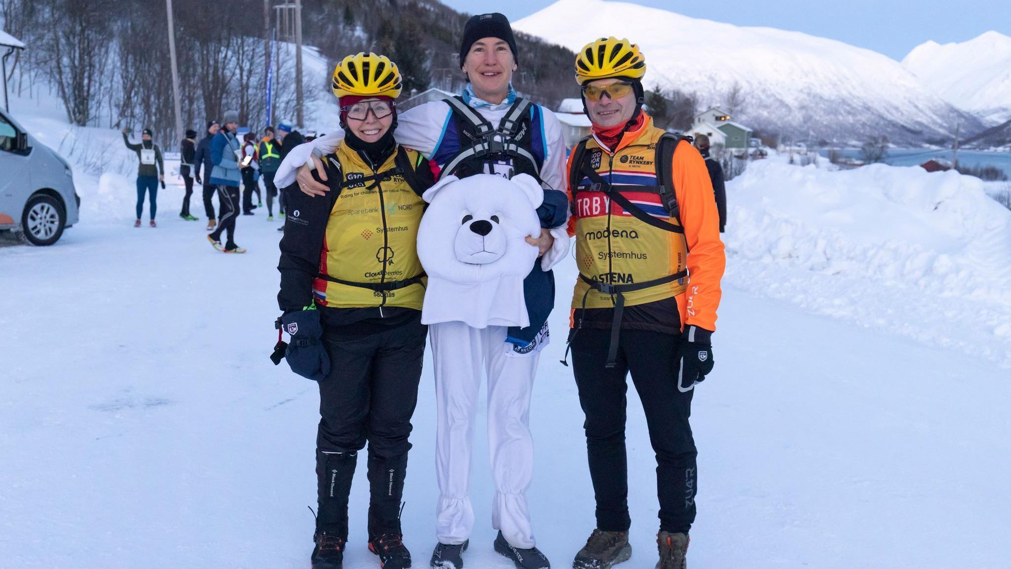 Three people standing on ice and snow in Norway. The woman is the middle is wearing a polar bear costume and holding the head, the other two people are dressed in orange and yellow. They are smiling at the camera. 