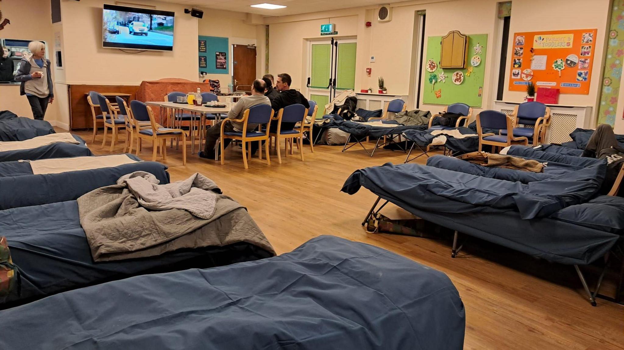 A woman standing and three other people sitting down. There is a line of beds with blue sheets. 