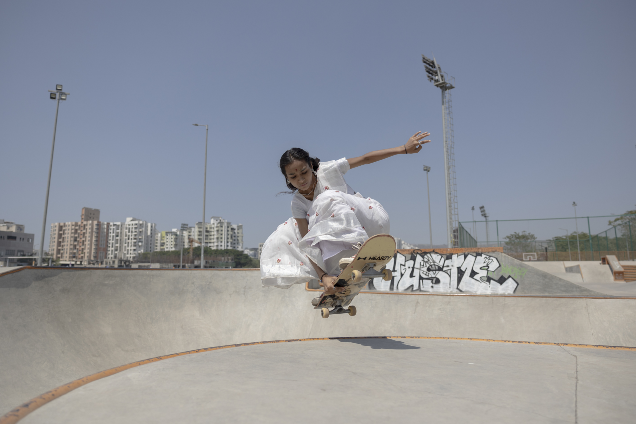 Shradda is leaping with her skateboard beneath her from a concrete skate park on a sunny day in an urban area, she is dressed in a white saree.
