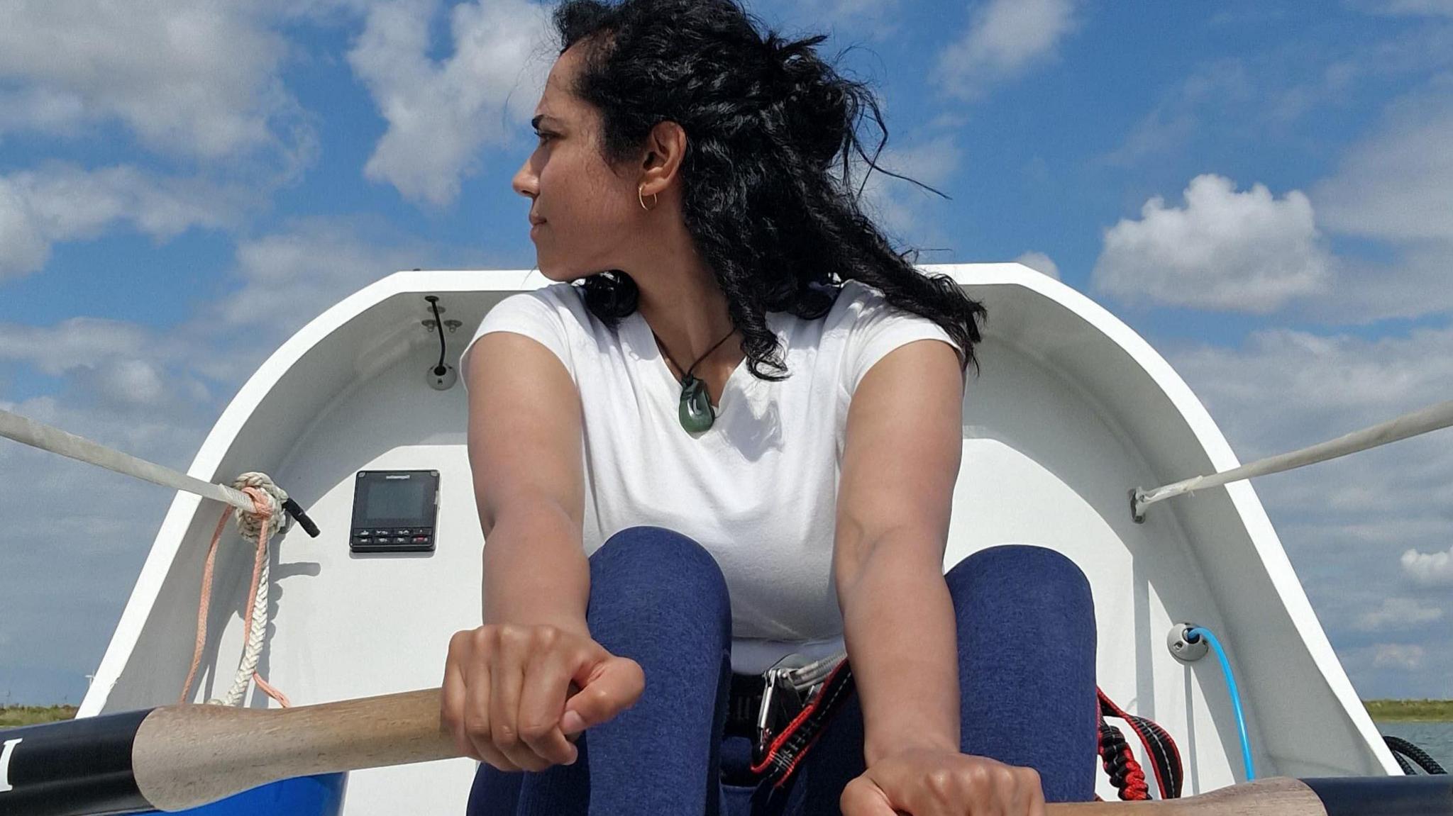 Ananya is seated on her boat holding the oars, gazing over her right shoulder at the horizon.