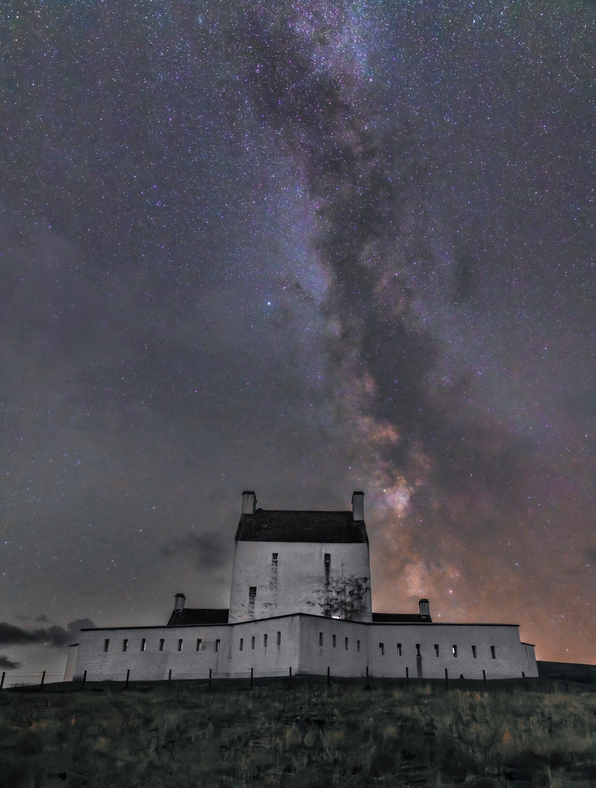A tall, white building with two smaller buildings on either end, surrounded by a white wall. The sky above is purple and pink and shows the stars in the Milky Way.