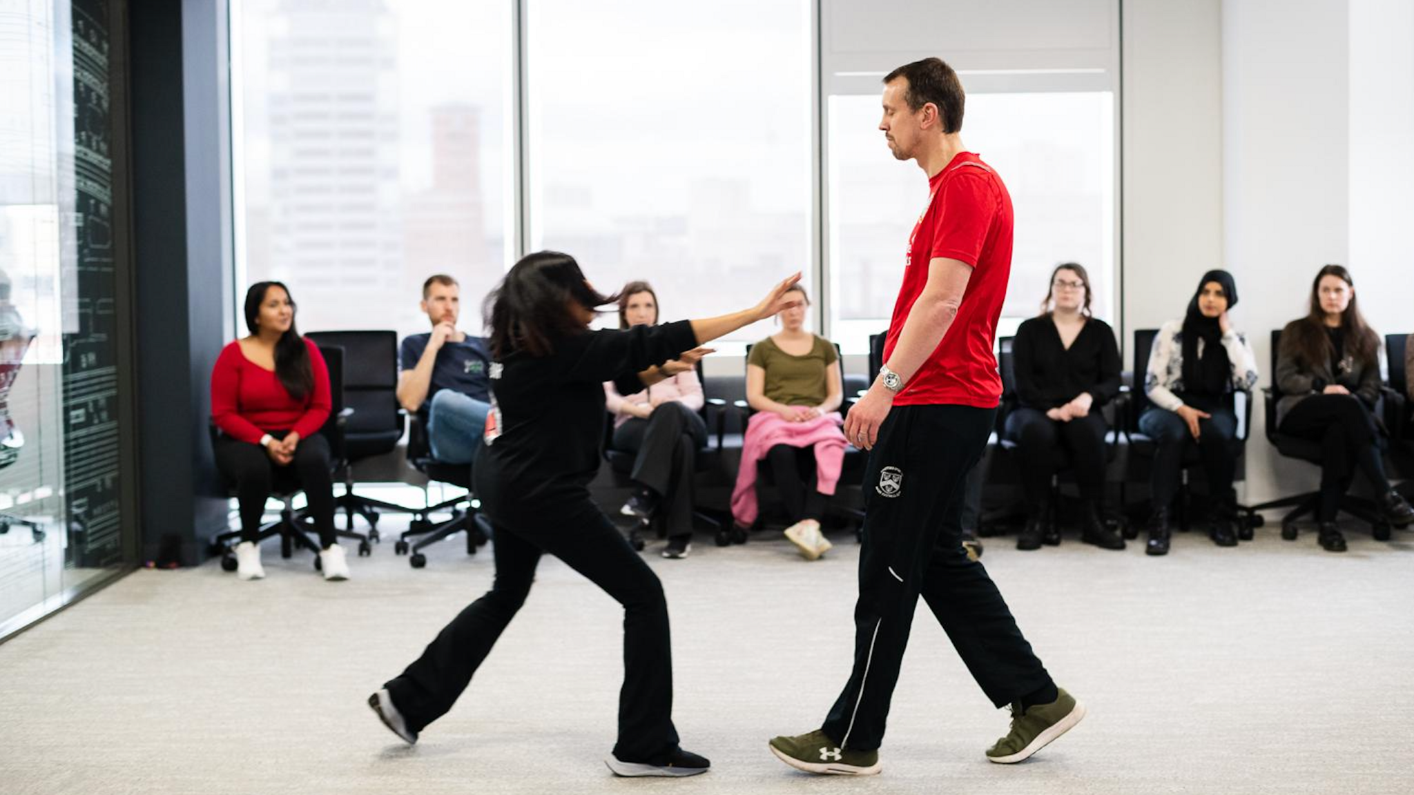 A woman is standing up demonstrating self defence in-front a group of other women who are sitting down and watching