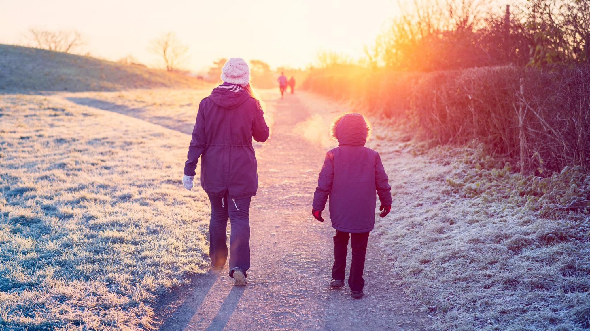 A woman and young boy wearing winter wear walking on a path with frosted grass around them. 