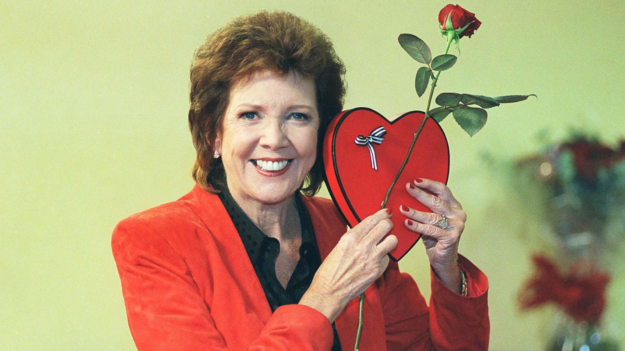 Cilla Black smiling and wearing a red jacket holding a red heart-shaped box and a red rose