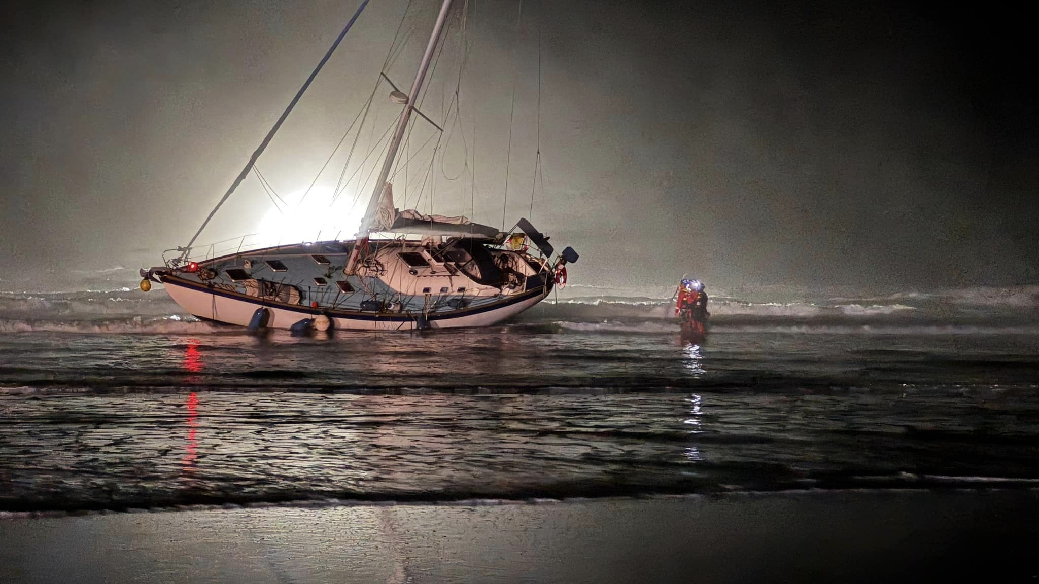 An overturned white sailing yacht on a flat sandy beach beach at night, illuminated from behind. The mast - without sails - is pointing about 35 degrees, towards land. The tide is half out, and a person in red stands halfway between the shore and boat ankle-deep in water.