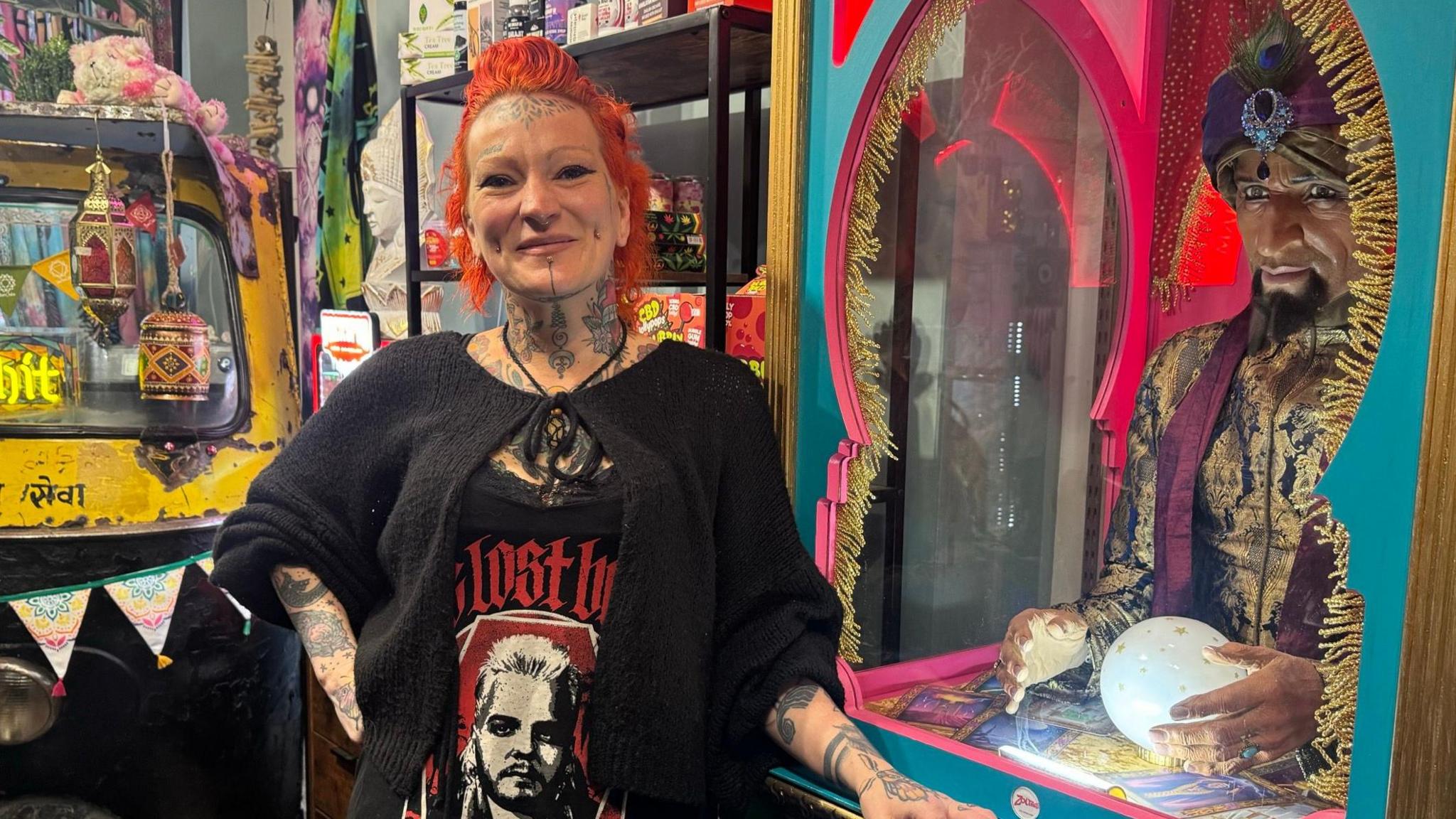 A tattooed woman with orange hair, wearing a Lost Boys dress, stands next to a fortune telling machine.