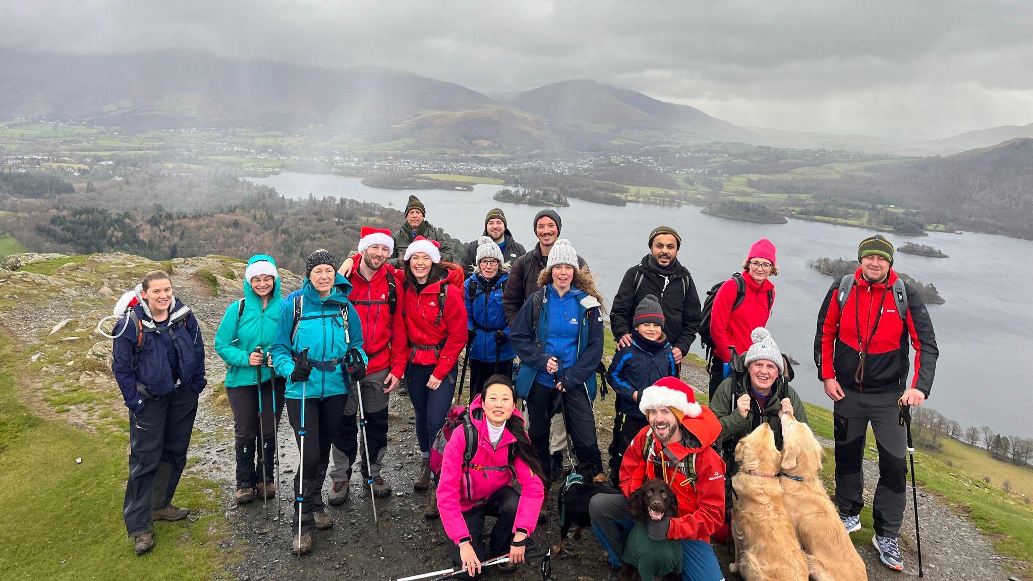 A group of people, some with Christmas hats, are on a path above a Lake District lake.