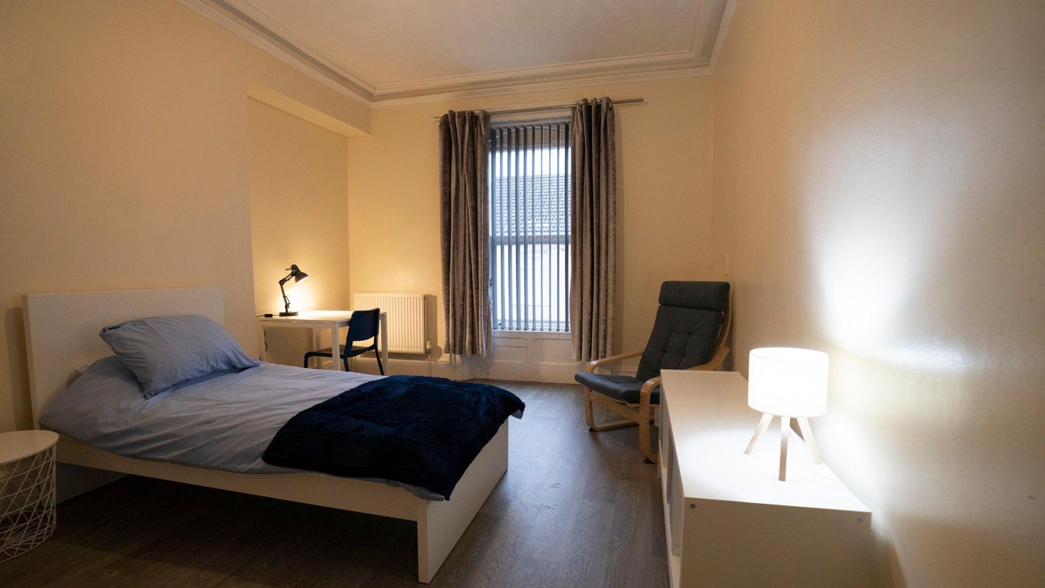 A bedroom at Teesdale House. It is painted in an off-white colour and has brown laminate flooring with a long window with blinds and curtains. There is a bed with a bedside table to the left and desk and a chair to the right. Opposite the bed are an armchair and a storage shelf. There are lamps on the shelf and the desk.