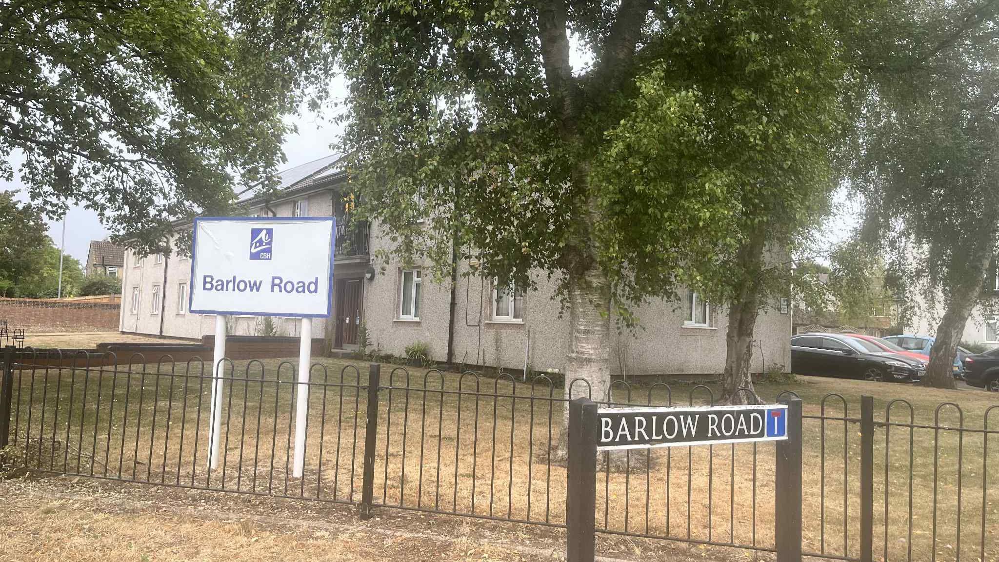 Barlow Road, as seen with two distinct street signs—one in blue and white on white posts, and another in black with white lettering—set against a backdrop of fencing, trees, and a nearby building.