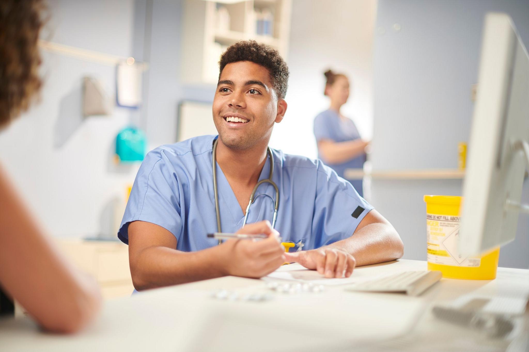 A member of hospital staff with a stethoscope sitting at a computer in a clinical setting and holding a pen 