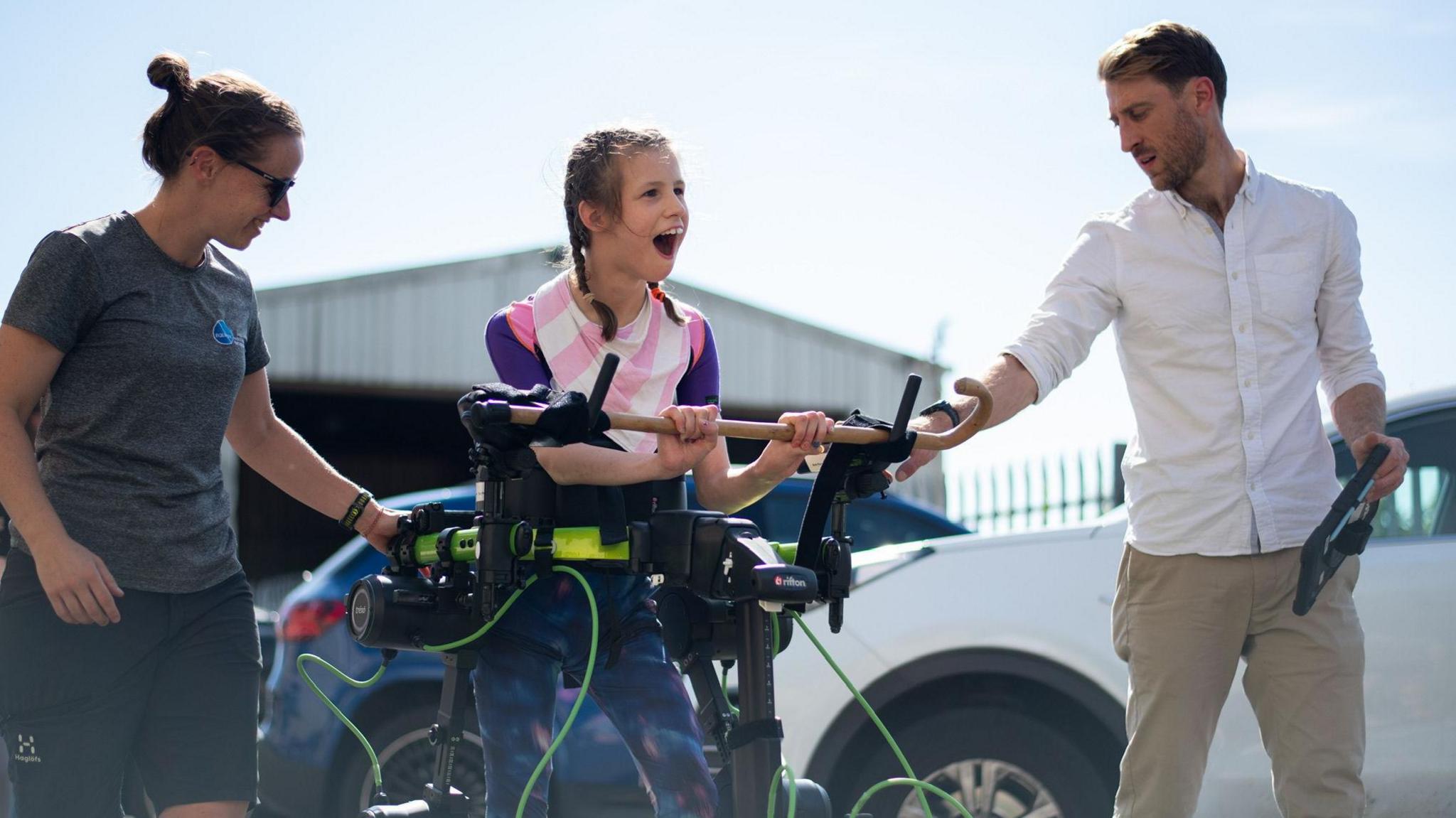 Brook stands, holding onto a horizontal wooden bar. The exoskeleton is black and runs down her legs, with a green bar around her middle. Brooke, whose dark hair is tied in pigtail plaits, smiles, and has a man standing on one side of her and a woman on the other. 