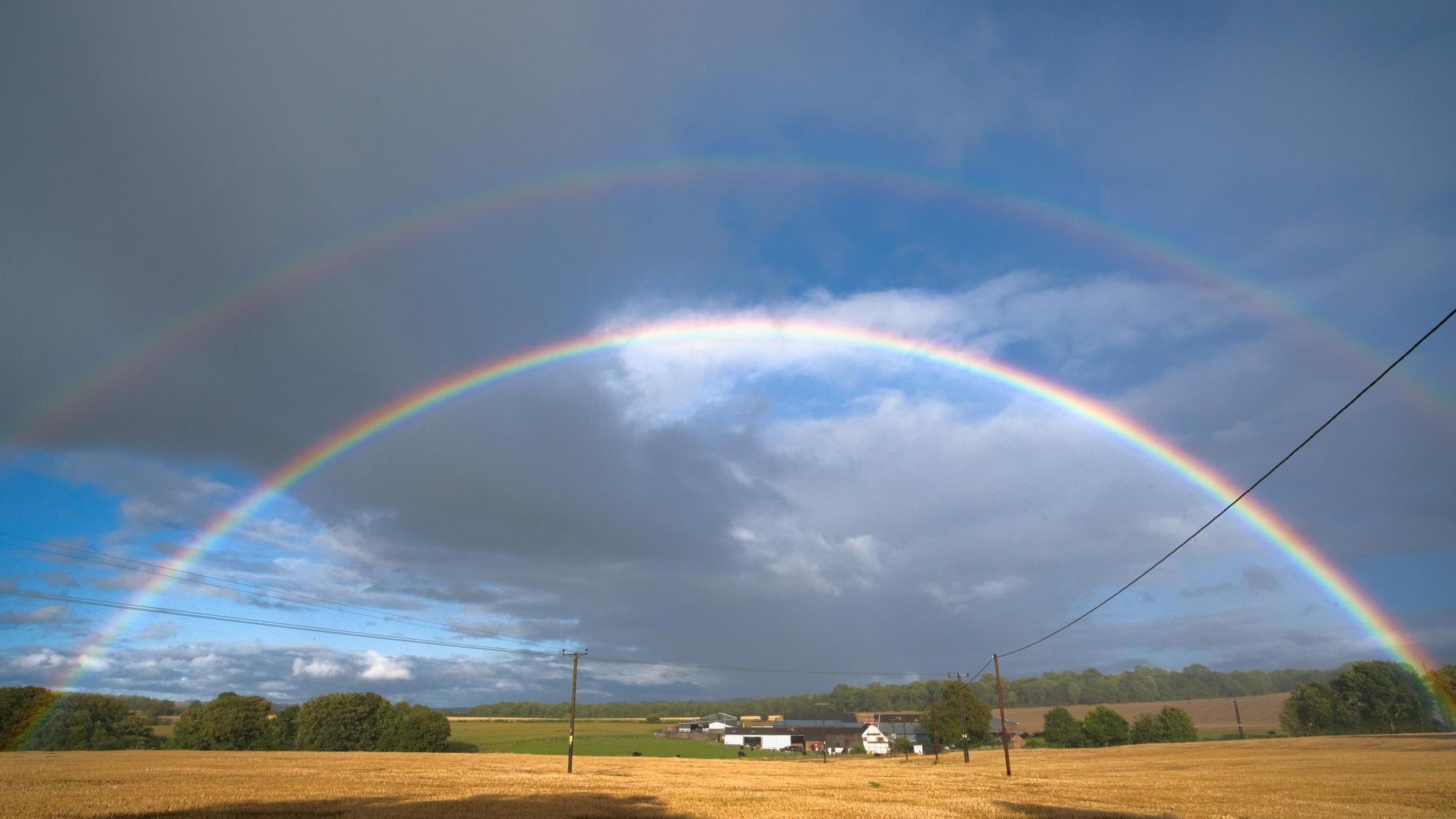 A rainbow clearly arches across the landscape, which is a golden field, a green one, farm buildings in the distance. Above the clear rainbow is a slightly more faint one.