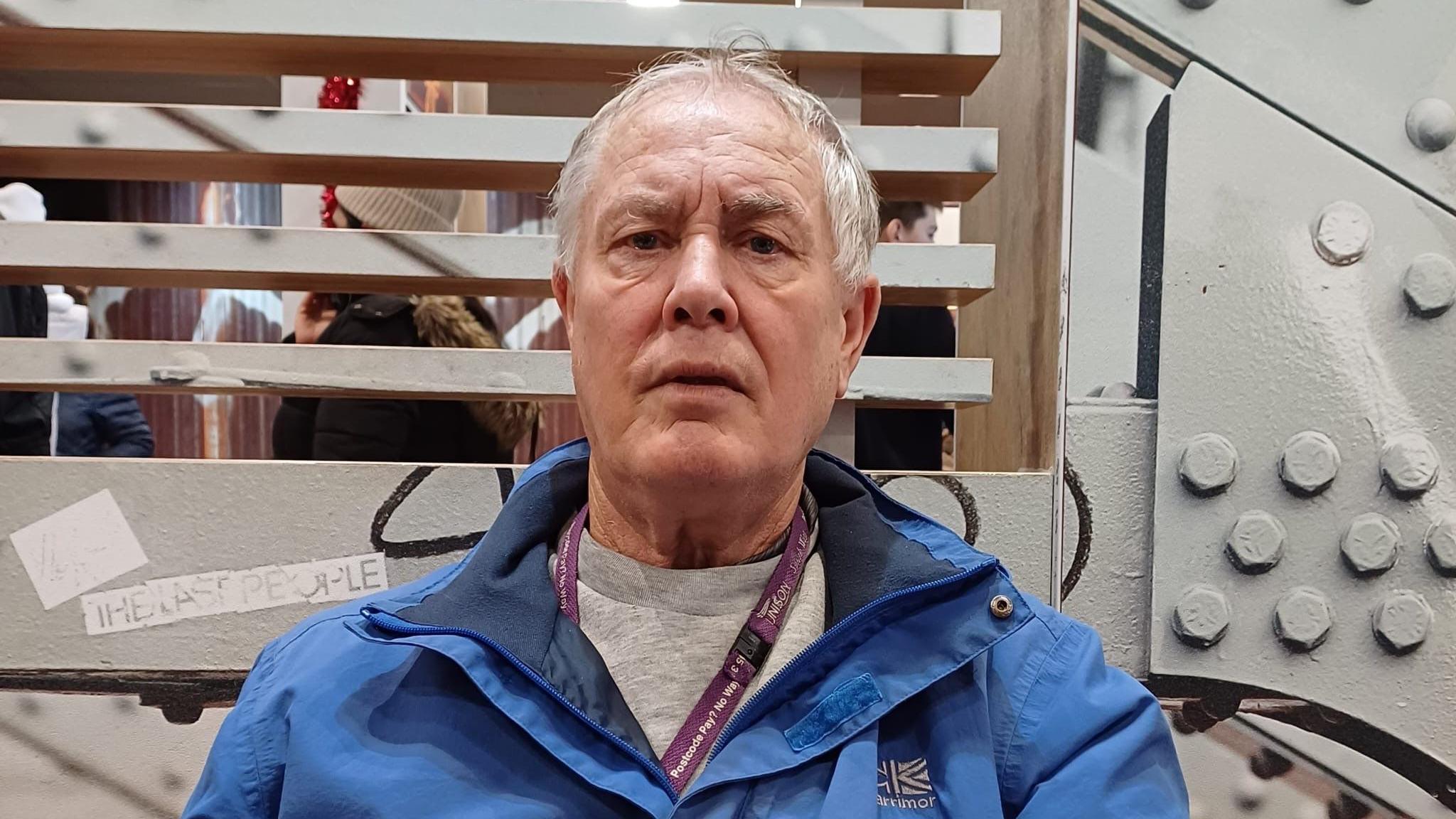 Stephen is looking at the camera with a neutral expression. He's wearing a grey jumper and blue coat, with a purple landyard around his neck.Behind him a grey industrial looking wall.