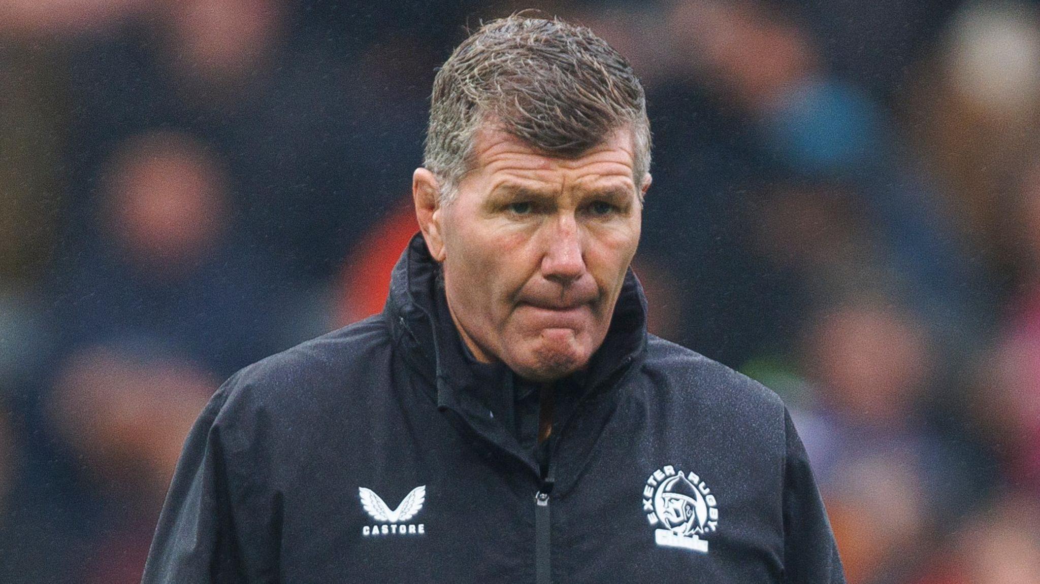 Rob Baxter, wearing an Exeter Chiefs waterproof jacket, watching his team warm-up on a rainy day