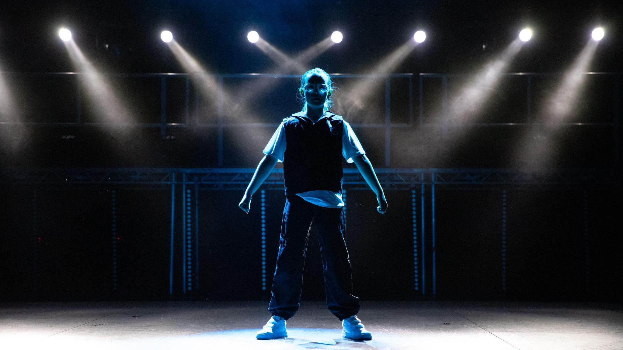 A young dancer stands on a stage. They are in the shadows of the lighting, and there are blue lights surrounding them. They are standing with their legs apart and their arms down, with their hands in fists. They are wearing glasses.