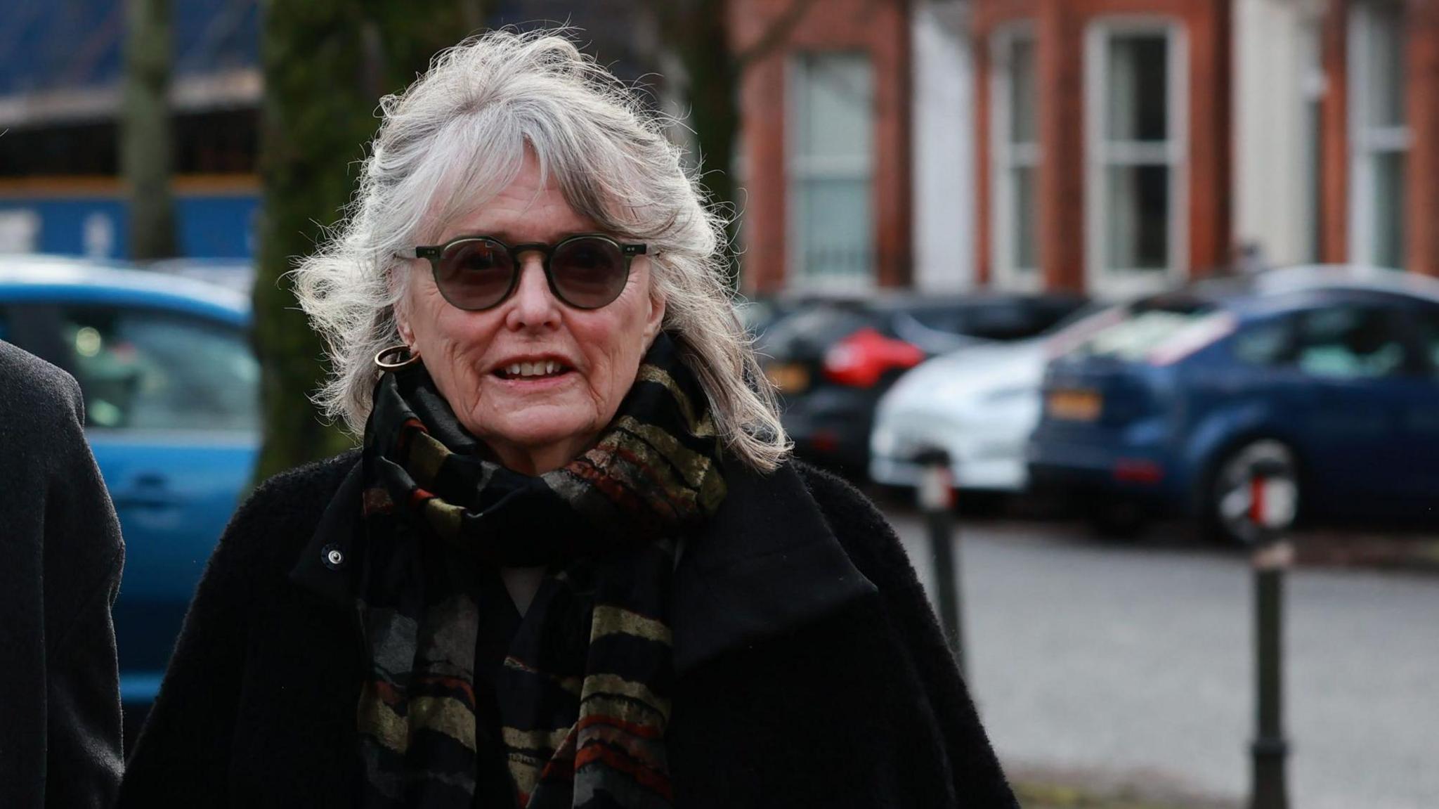 Marie has white hair, shoulder length. She has brown sunglasses and hoop earrings. She is wearing a black coat and colourful scarf, yellow and black tones. 