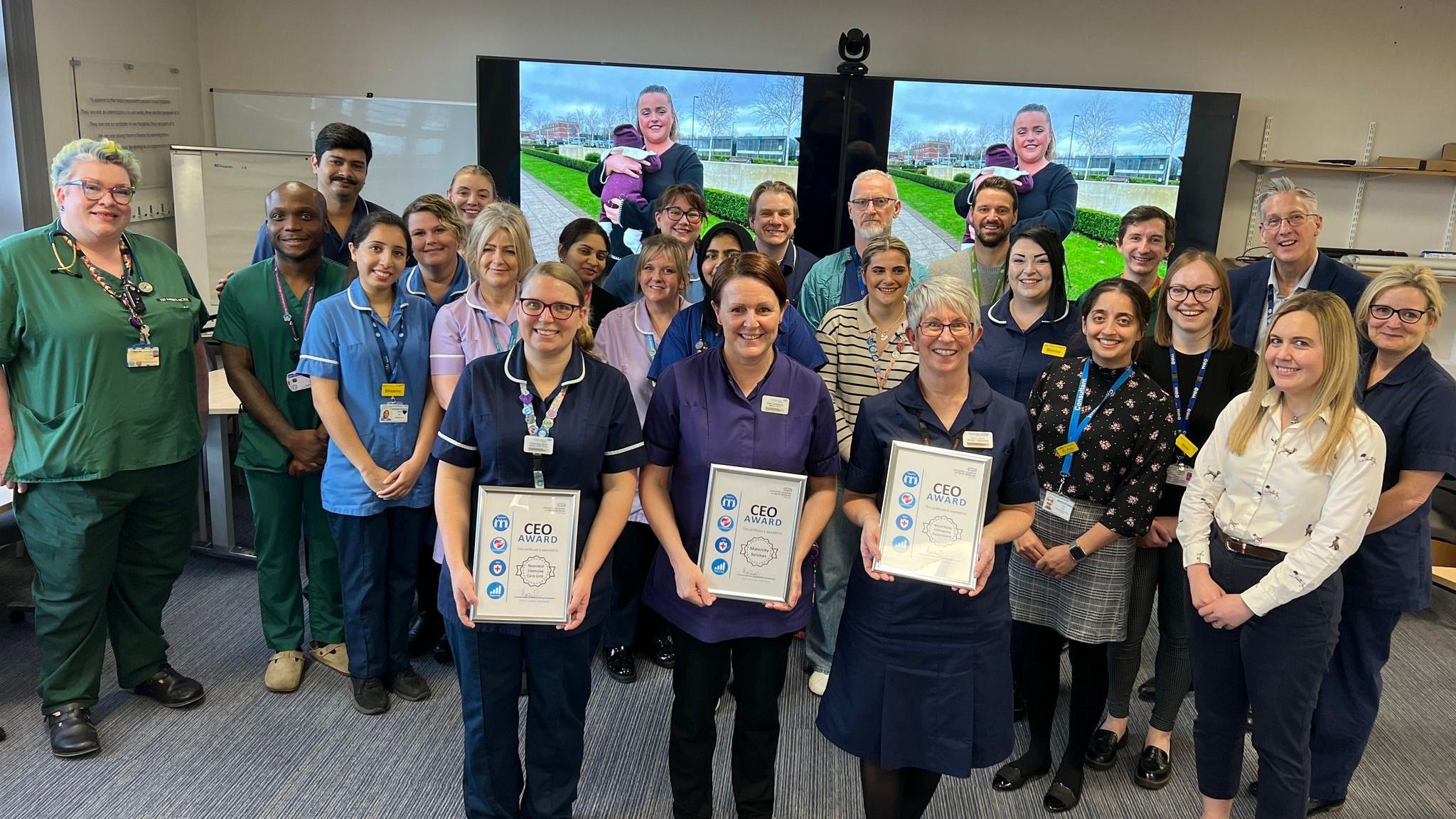 A team of people - some wearing medical scrubs - stood in a group, smiling while posing for the camera. Three of the group, stood at the front, are holding framed certificates.