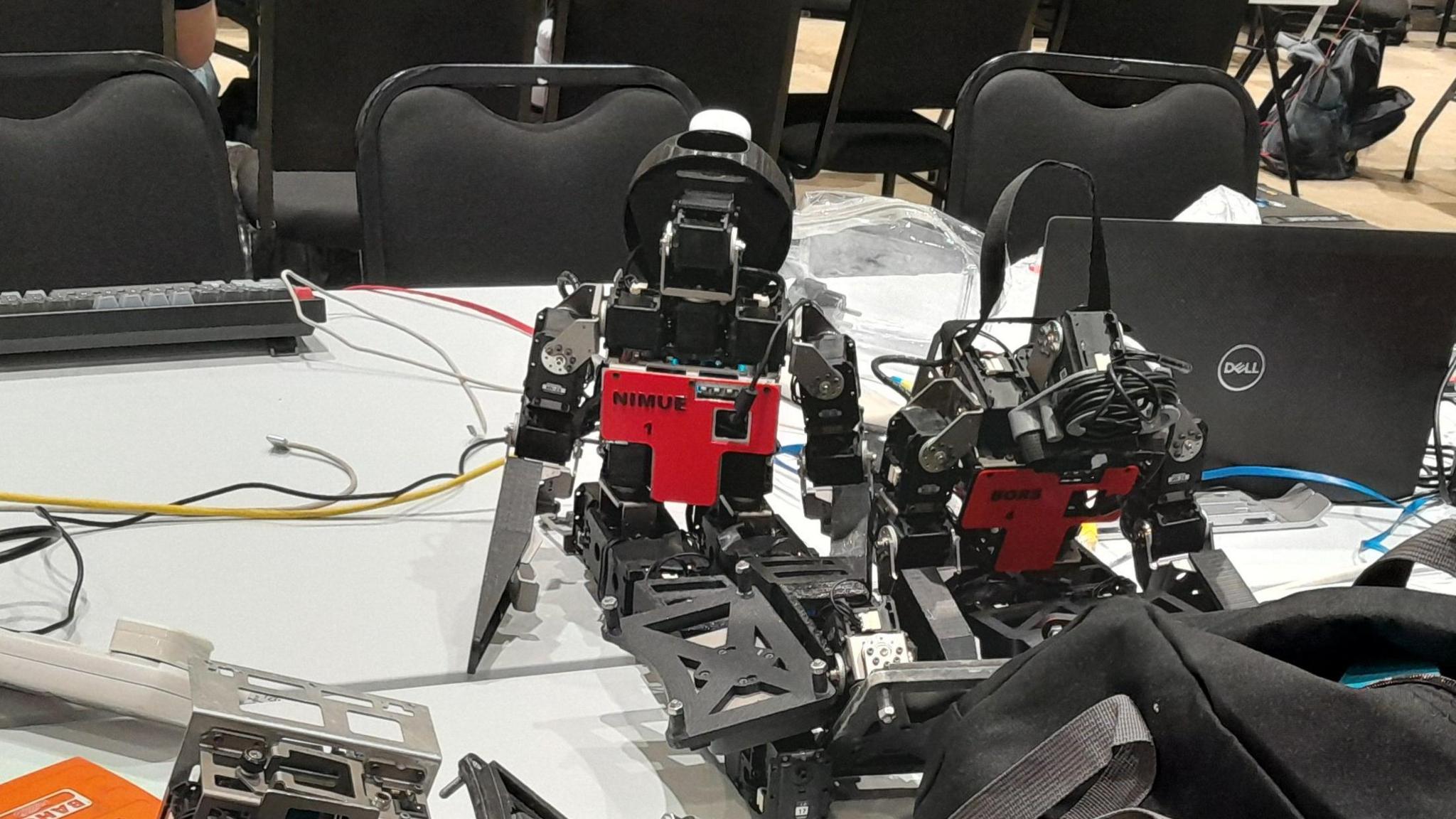 A small robot on a desk surrounded by other equipment including a computer and a keyboard. There are chairs in the background.