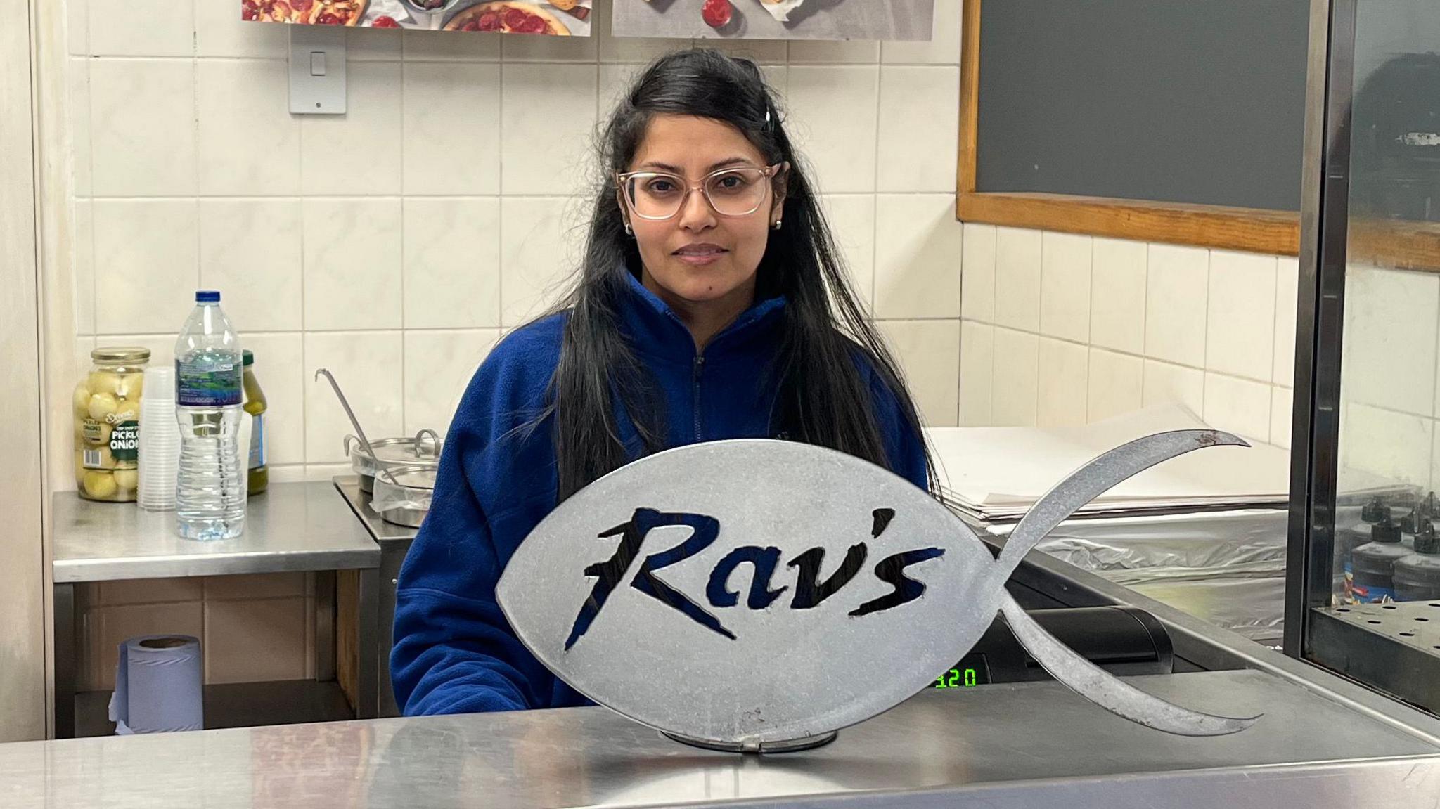 Rav stands in front of the fish and chip shop counter. There is a silver sign in the shape of a fish saying Ravs. She has long black hair, wears glasses and is wearing a blue fleece top. 