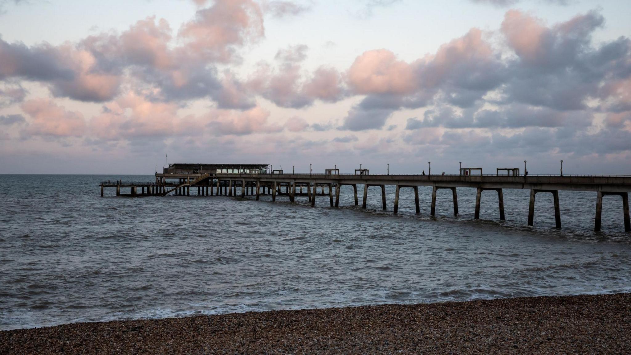 Choppy waters surround Deal Pier, a wooden structure - with a strip of pebbly beach in the foreground. 