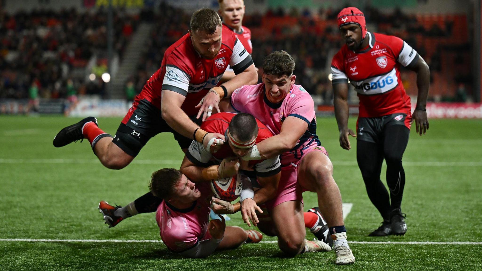 A group of players commit to a tackle as Gloucester play Exeter in the Premiership Rugby Cup at Kingsholm. A Gloucester player is going to ground with the ball in his hands, while two Exeter players tackle him, one of them grimacing with the effort. In the background other Gloucester players look on. Gloucester are in their traditional colours of red and white while the Exeter players are in their change kit of light pink and black