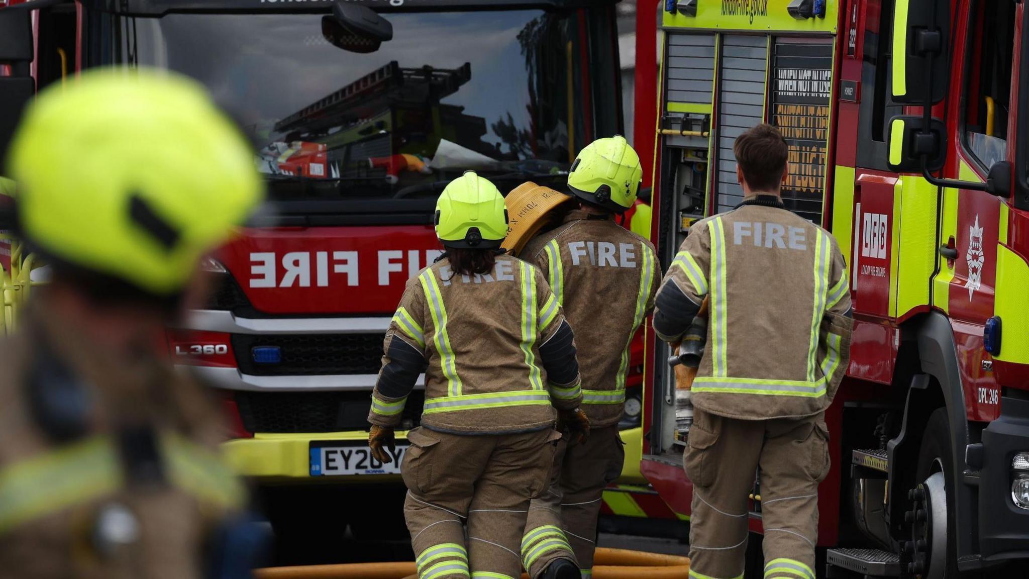 Three firefighters walk towards a fire engine with another crew member blurred in the foreground. They wear brown uniforms with fluorescent strips, with 'fire' printed on the uniforms in big white letters.