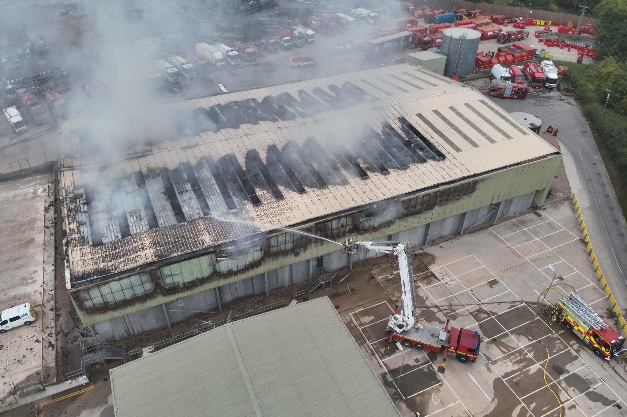 An aerial view of the fire at the waste depot. A large rectangular building has got blackened, charred areas and there is smoke billowing from the roof. Two fire engines are parked around the front of the depot, with one having an aerial platform which is hosing water onto the roof of the burning building.