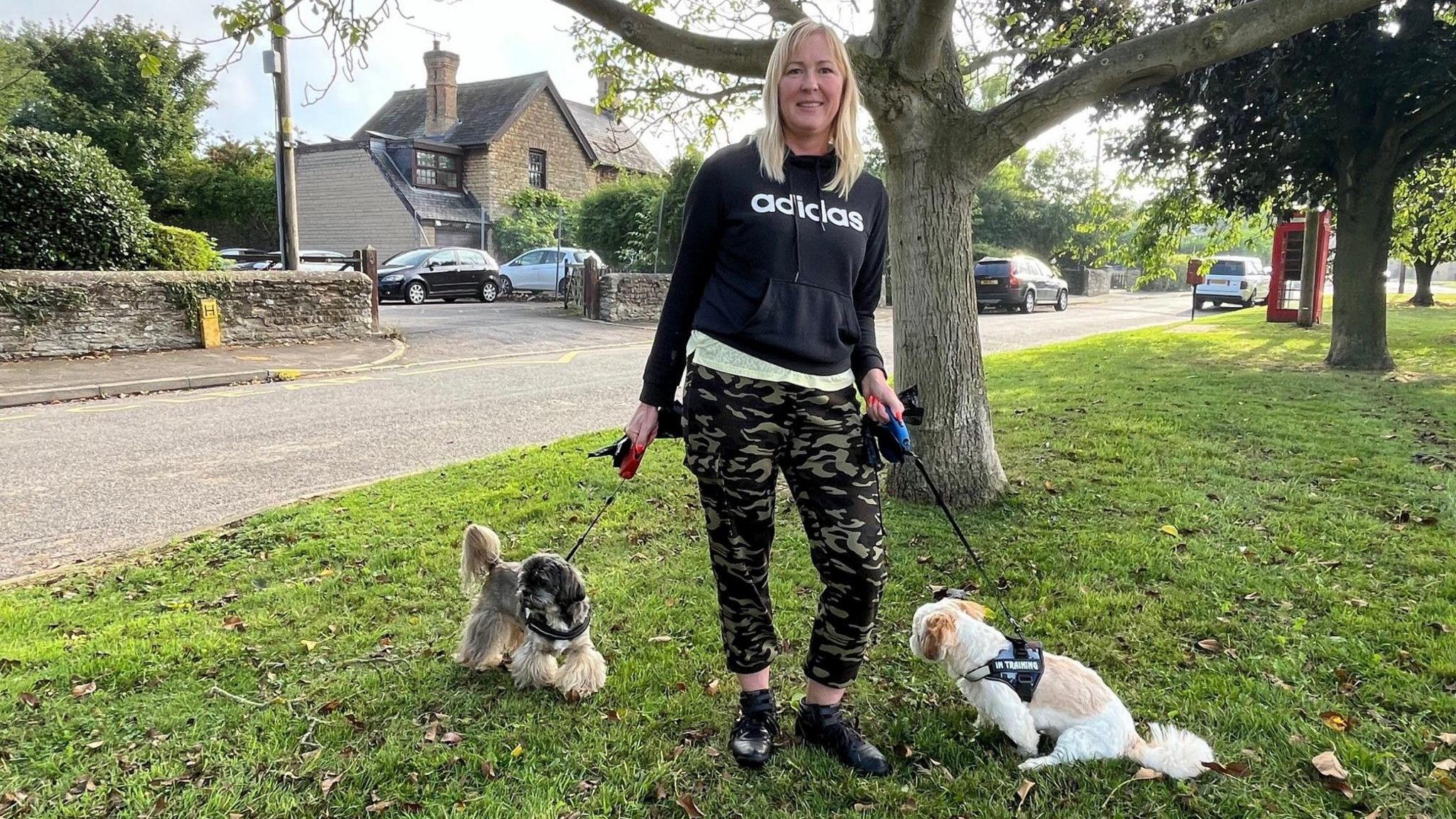 Simone Hunt, with blonde shoulder-length hair, wearing a black long-sleeved top with a white Adidas logo and green and brown camouflaged trousers, pictured with two small dogs on leads. 