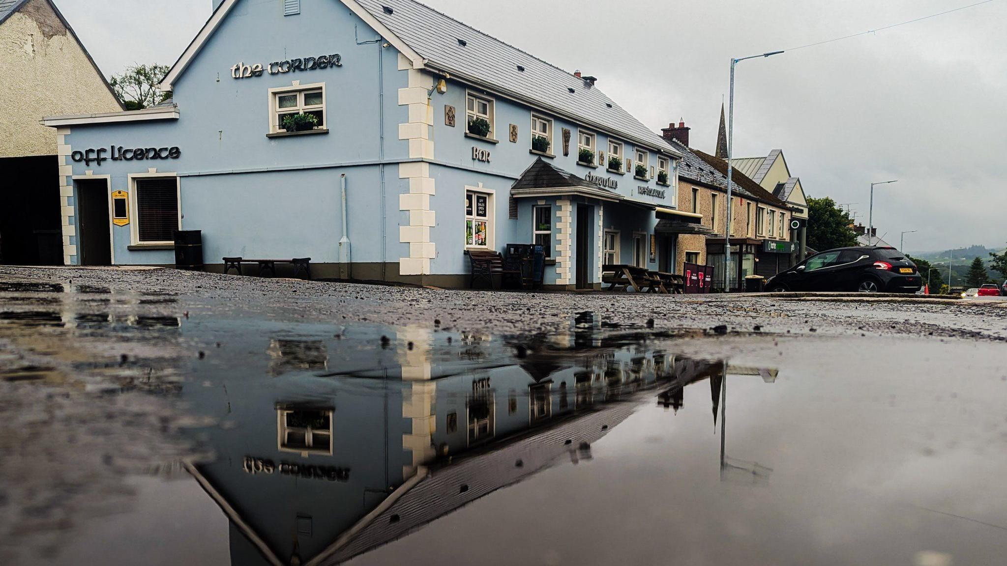 A large puddle on the ground outside a blue building. The building is in the reflection of the puddle.