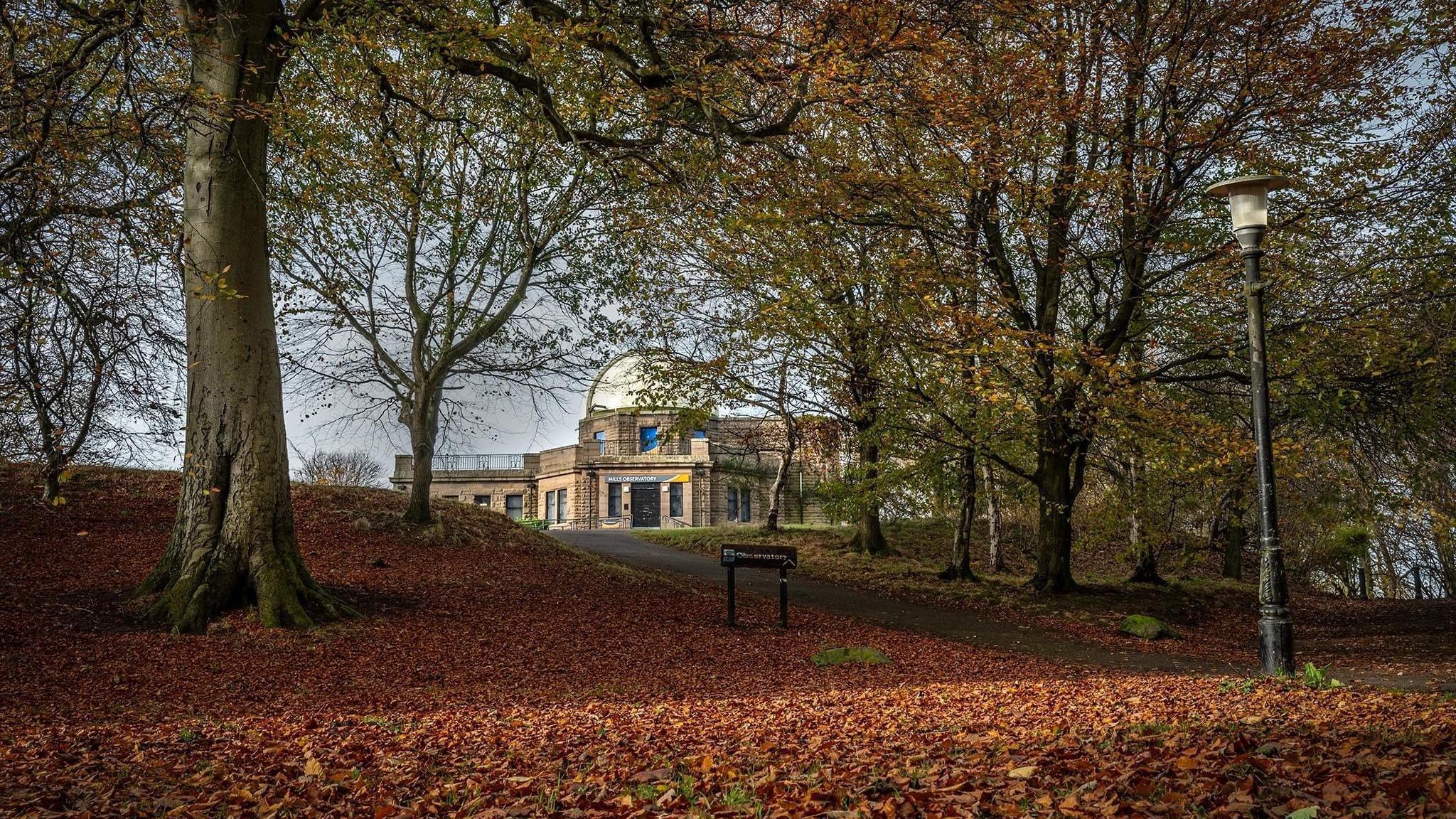 An observatory building is viewed through a gap in autumnal trees which have shed many of their golden brown leaves across the grass in the foreground