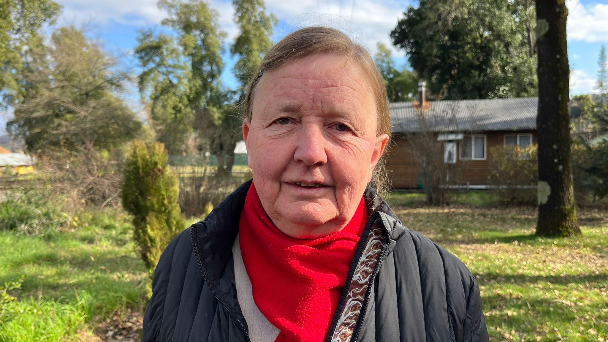 Erika Tymm, wearing a red scarf, looks at the camera while standing in Villa Baviera. Behind her a house can be seen and green grass and trees. 