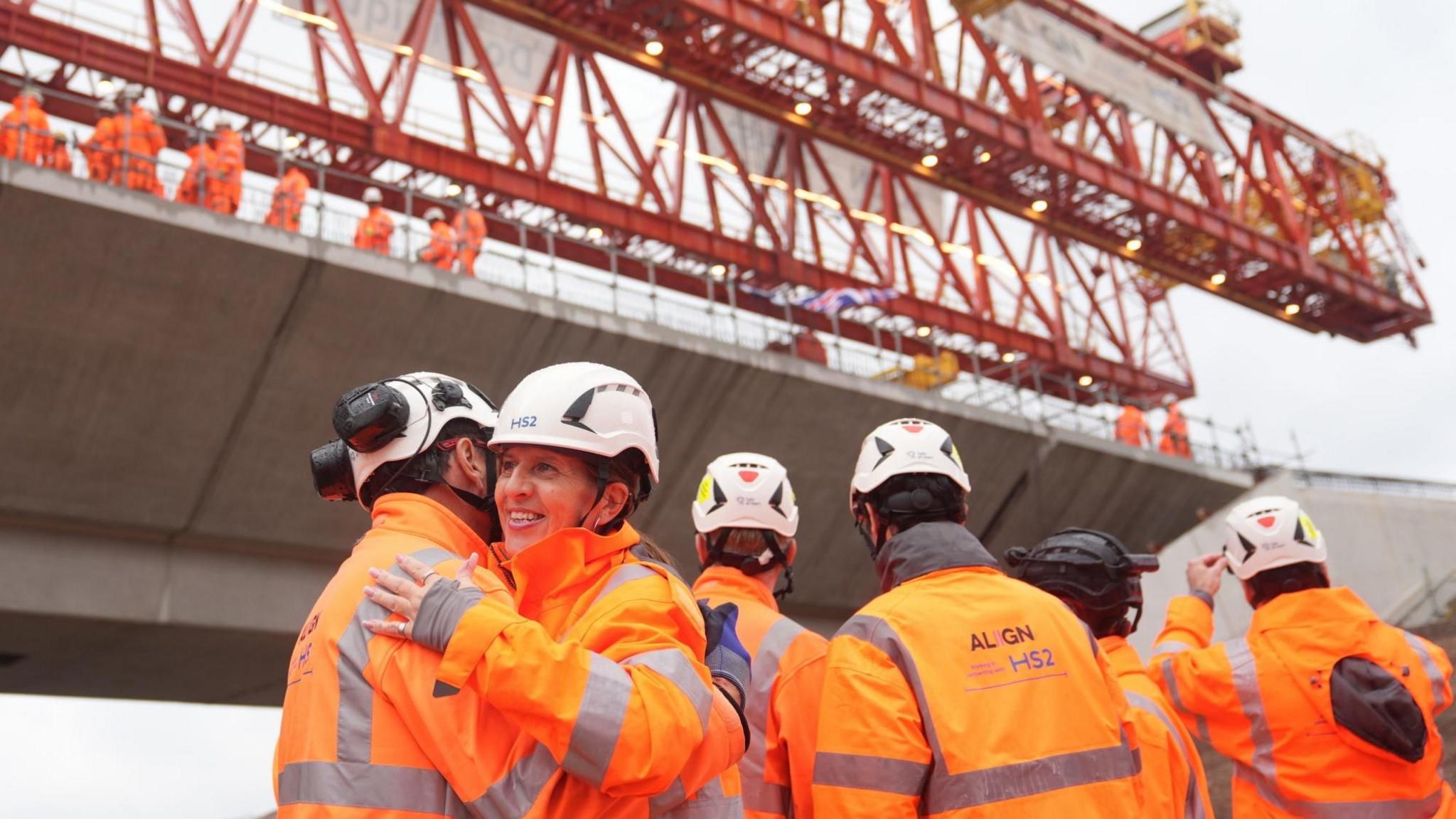 Men and women wearing orange high-vis outfits and helmet hug and smile near the newly completed bridge which is grey concrete.