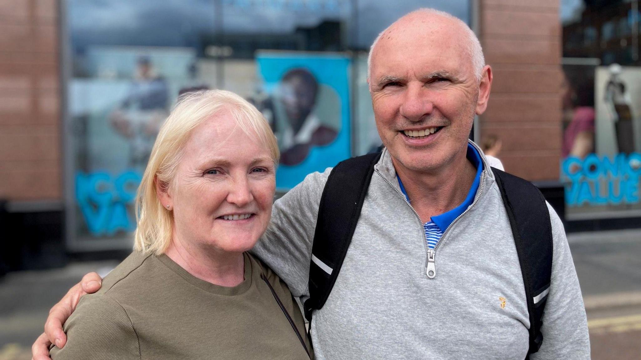 A woman and a man smile at the camera. The woman has blonde hair and is wearing a pale-green top. The man is wearing a grey top.