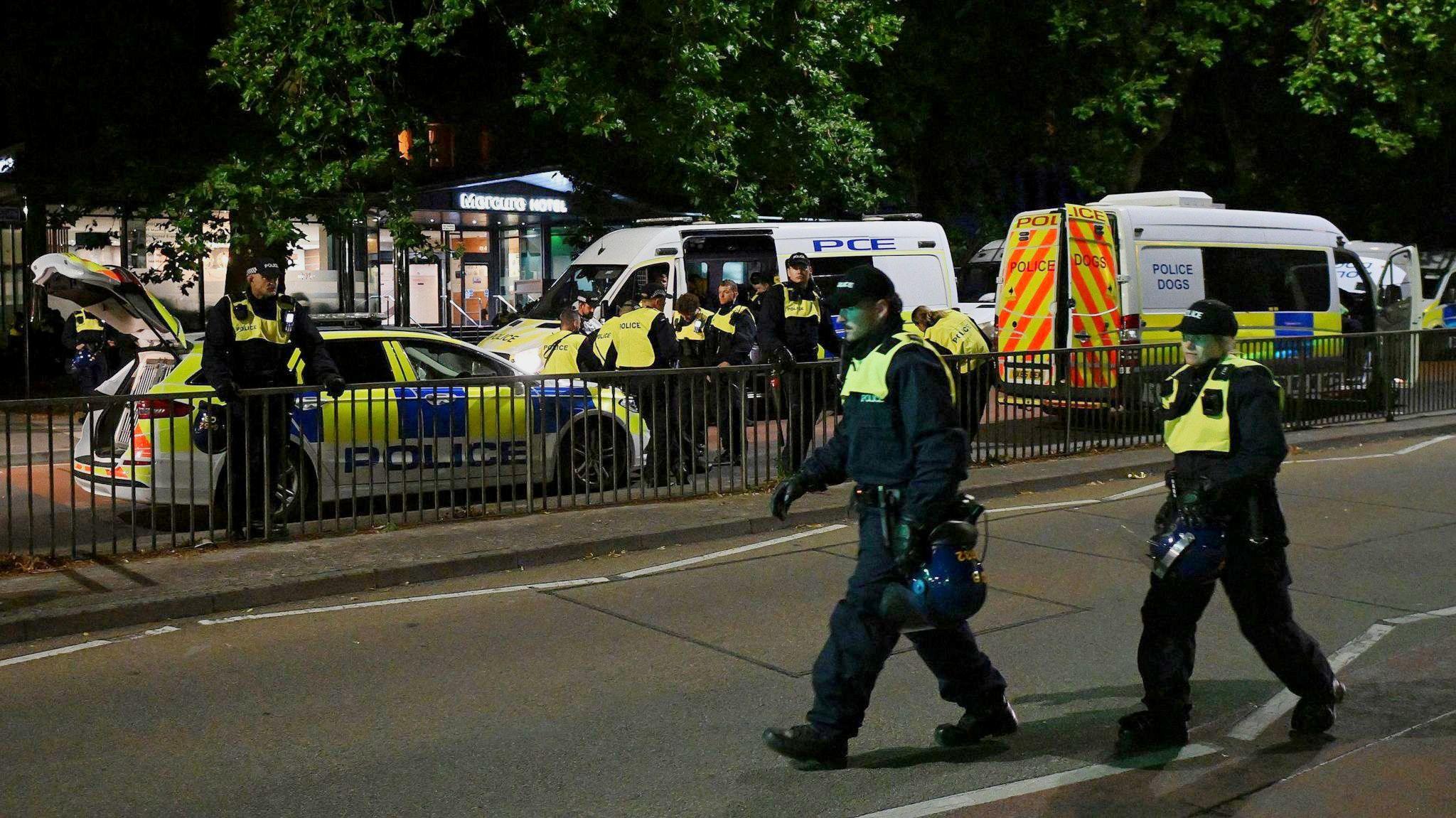 Two police officers in the forefront of the picture, carrying riot helmets in their hands. There are two police vans and one police care parked outside the Mercure hotel in Bristol at night, and approximately seven police officers are standing by them