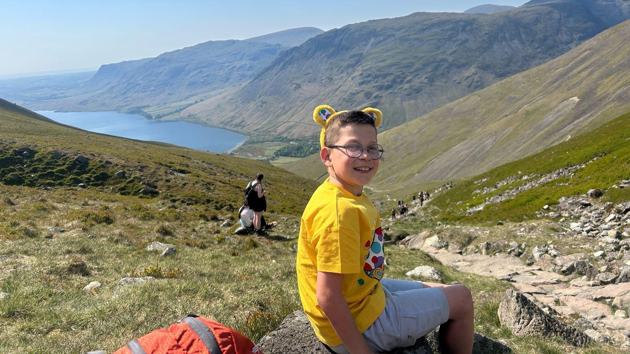 A boy with brown hair and glasses in a yellow T-shirt and white shorts sat on a rock, part way up a large hill which can be seen stretching down to a lake below and there are other large hills in the distance
