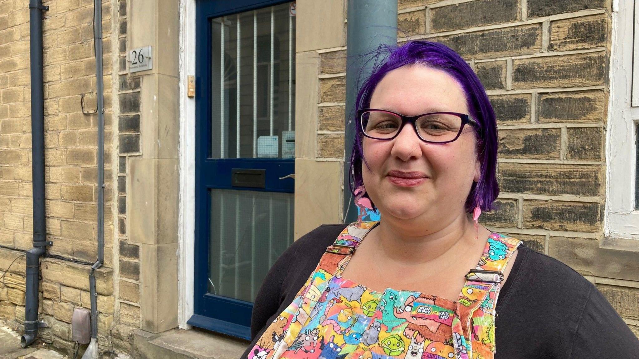 A woman with purple hair standing outside a dark blue door with glass panels and white security grilles.
