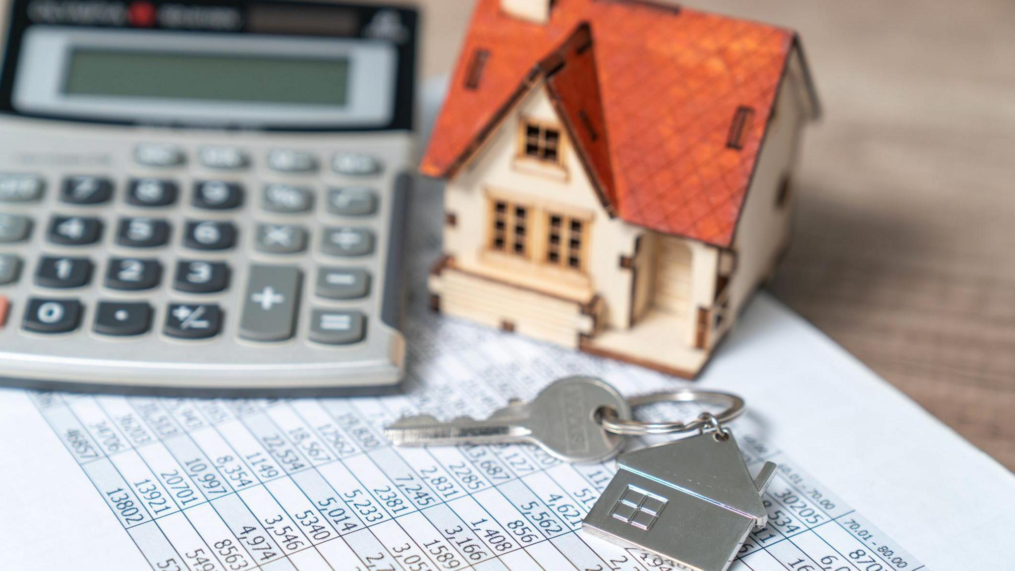 A small wooden home and a set of keys sat on top of a piece of paper which shows a table with lots of figures on. Beside them, there is a calculator